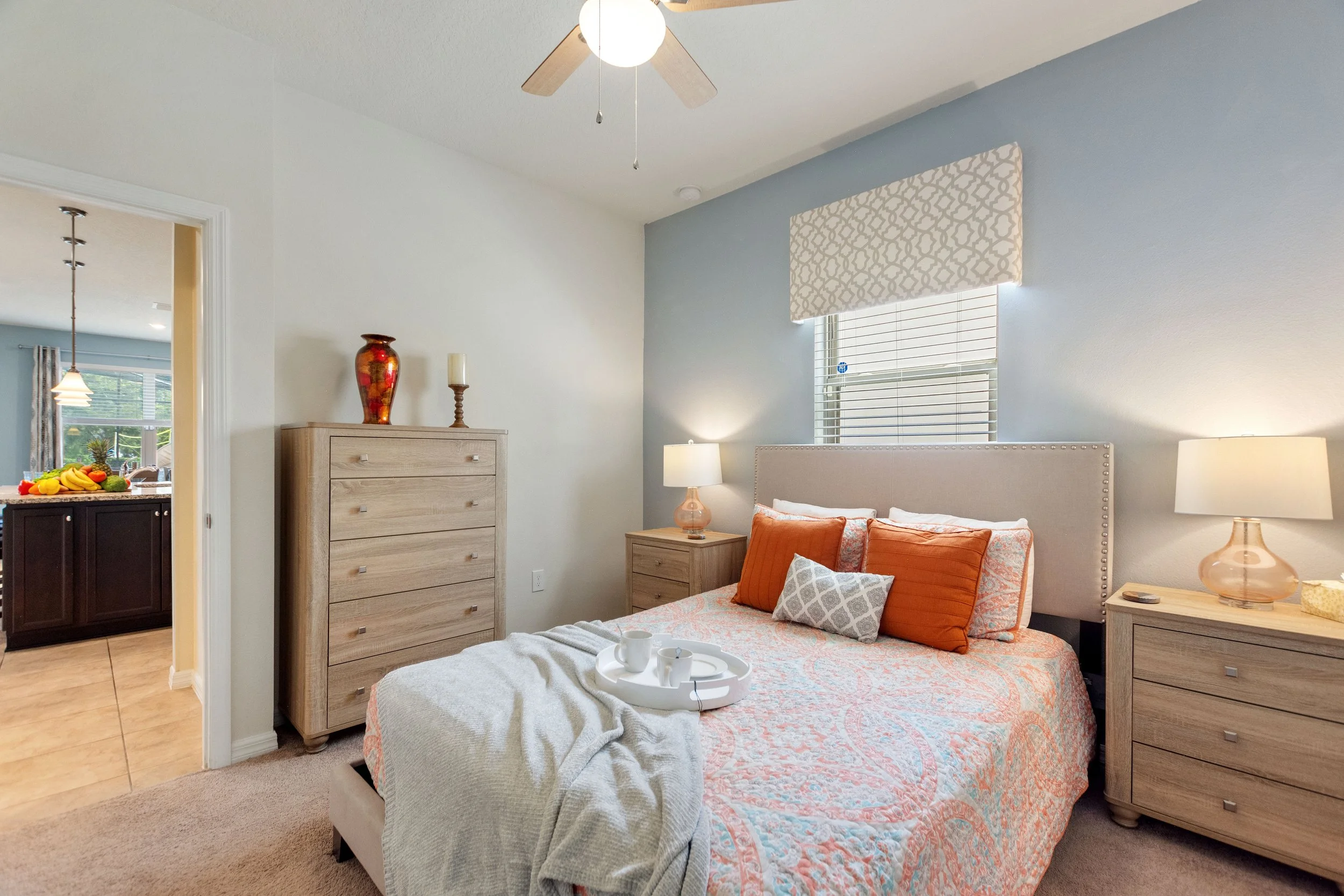 A cozy bedroom with a bed that has orange and patterned pillows, a beige headboard, and wooden nightstands with lamps. A tray with cups is on the bed. There are two dressers, one with a decorative vase and a candle. A window above the bed lets in nat
