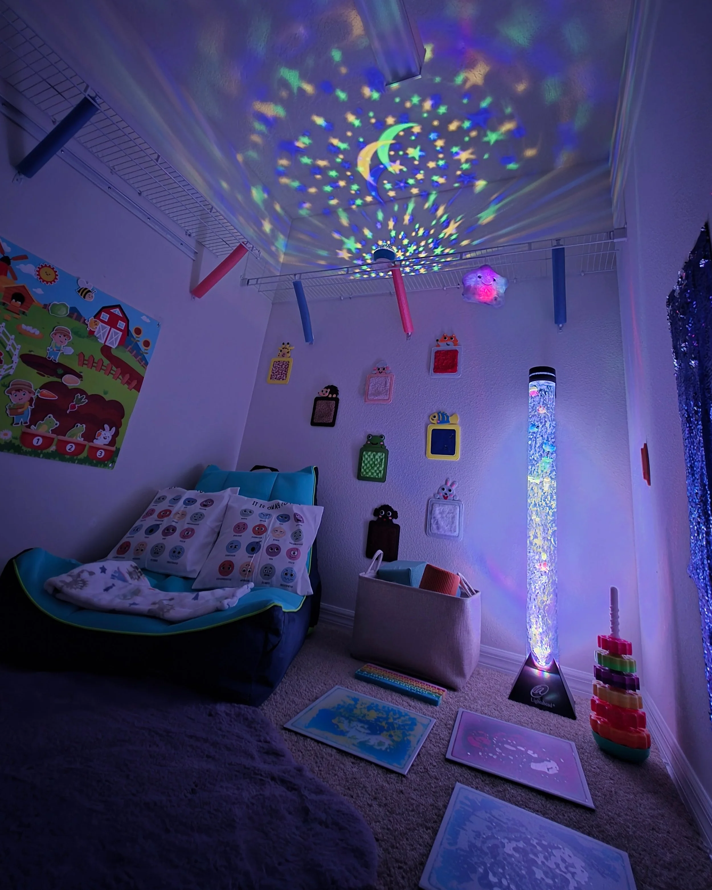 Children's bedroom with a ceiling projection of stars and moon. Small beds with pillows featuring emojis, wall decorated with colorful framed art, a tall star-shaped light, and a stack of storage bins.