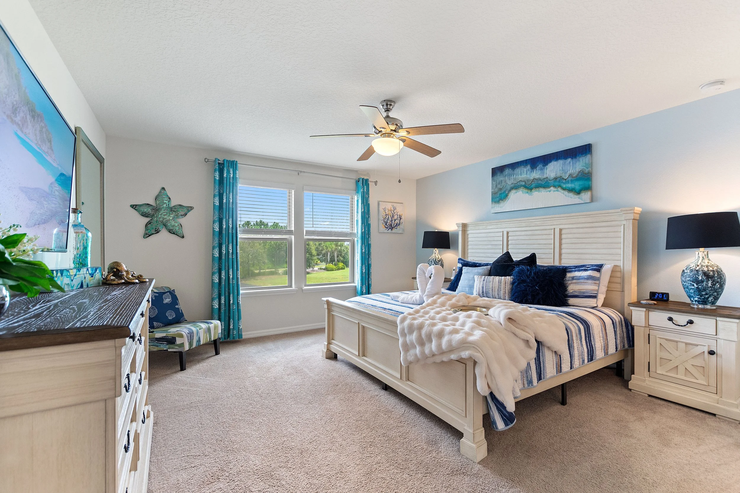 Bright bedroom with white furniture, blue and white striped bedding, navy pillows, and ocean-themed decor. Large window with blue curtains shows greenery outside. Ceiling fan with wooden blades. Decor includes wall art, a starfish-shaped decoration, 