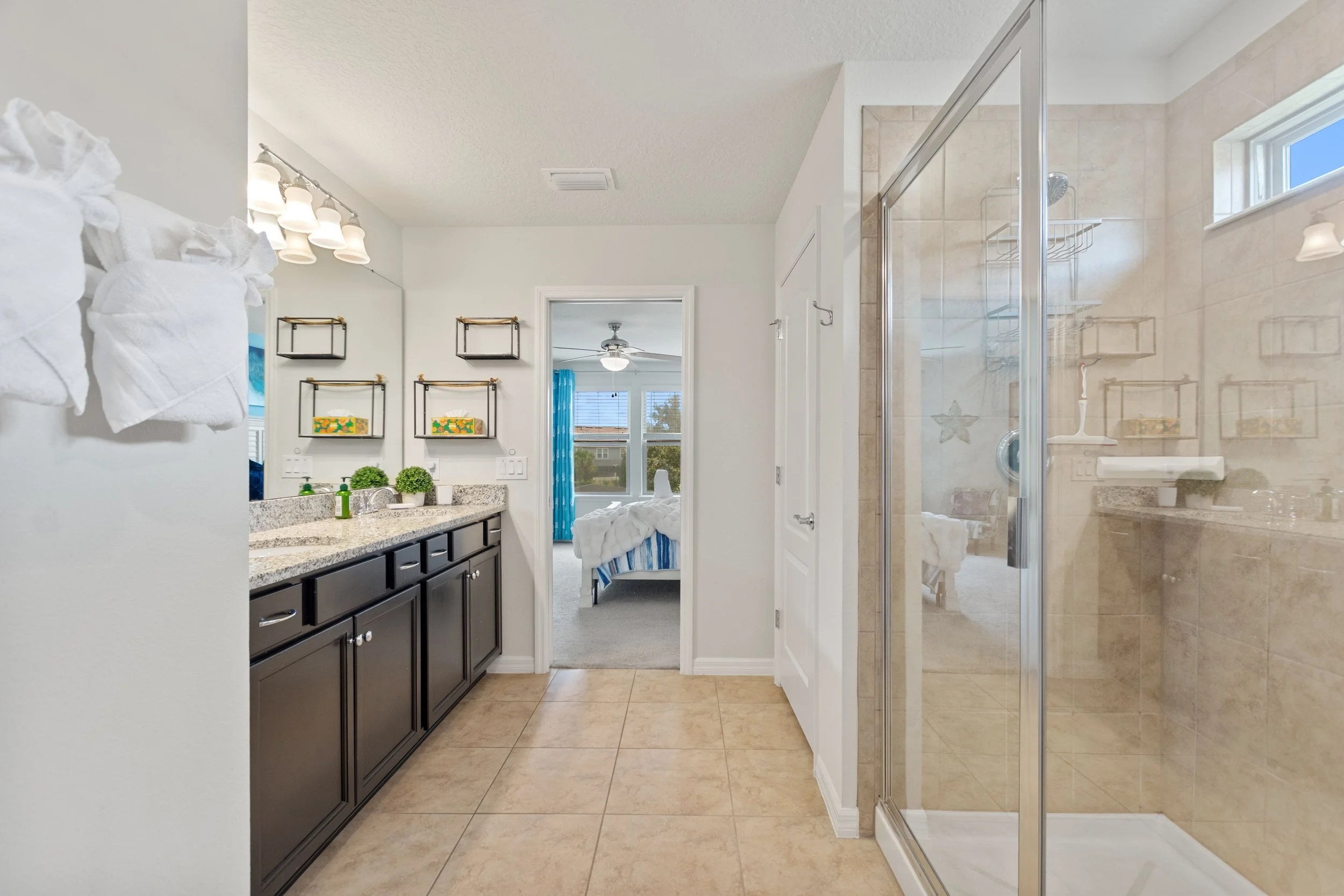 Bathroom with granite countertop vanity, large mirror, towel rack, and a walk-in shower with glass door, beige tiles, and a small window.