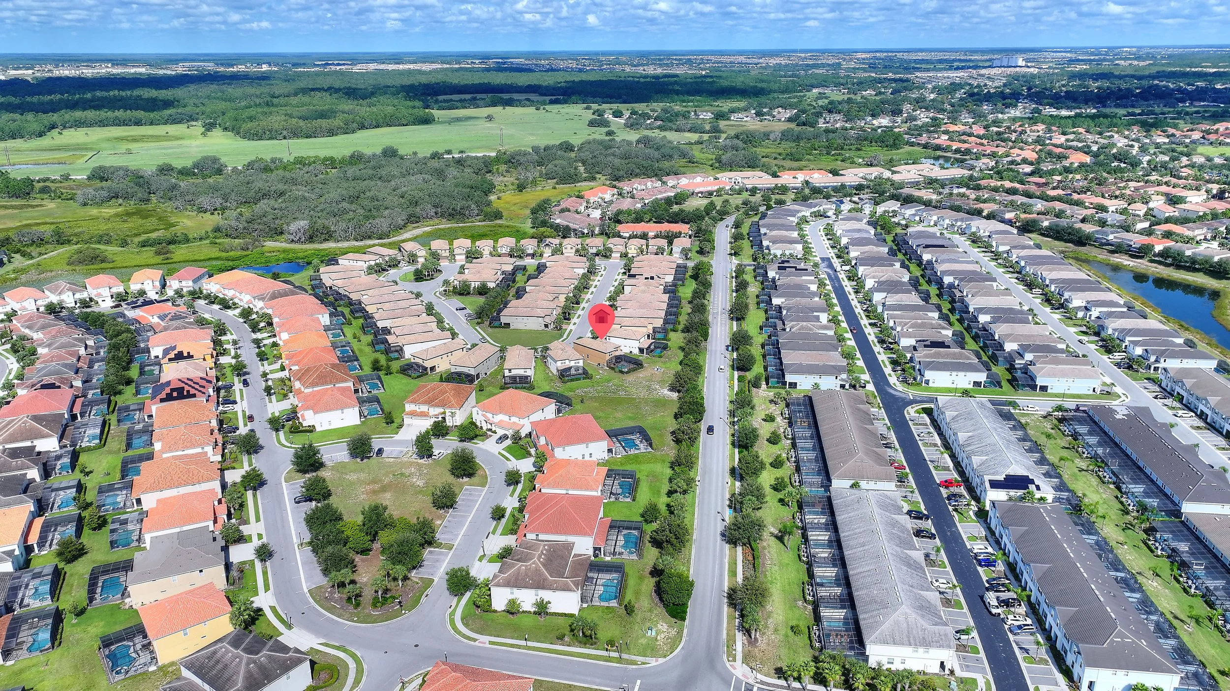 Aerial view of a residential neighborhood with rows of houses, some with swimming pools, green lawns, and trees. There is a canal or small water body on the right side, and open green fields in the background under a partly cloudy sky.