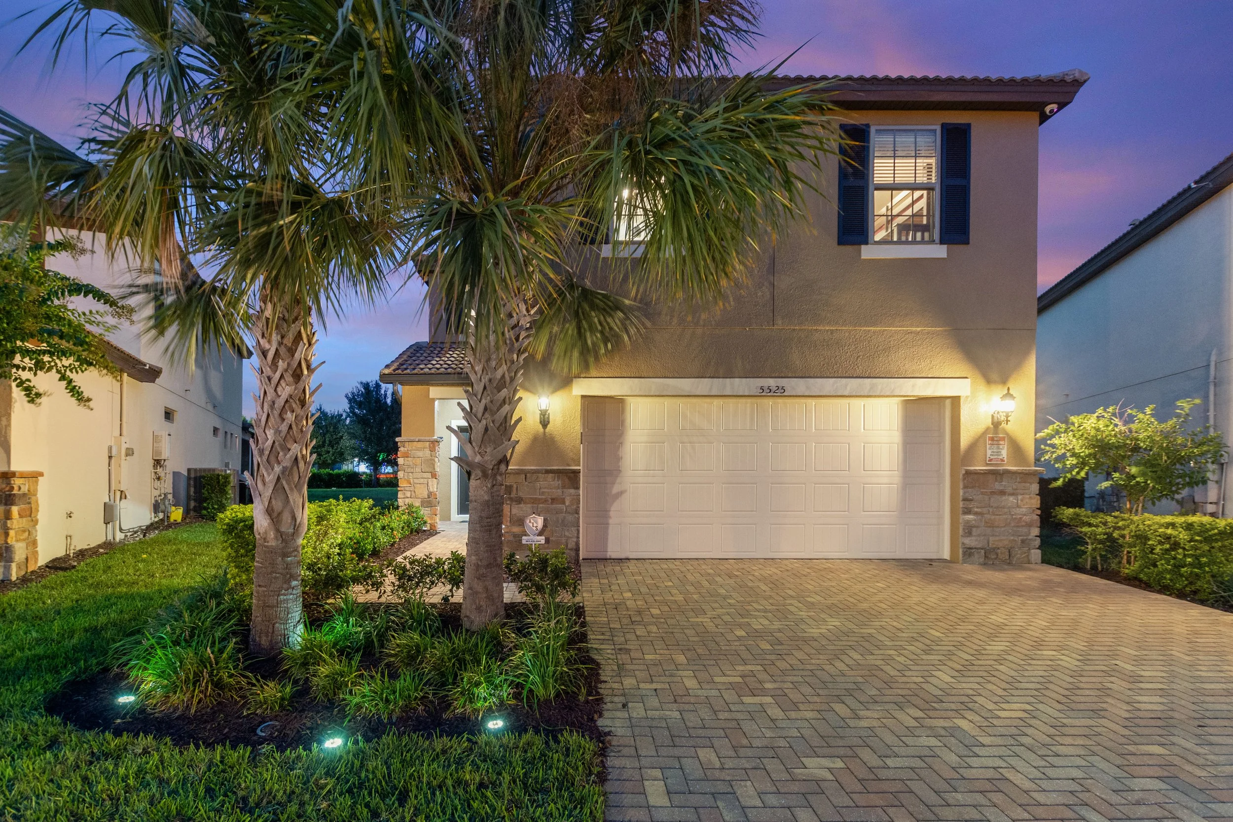 Two palm trees in a landscaped front yard with a paved driveway leading to a two-car garage, illuminated by exterior wall lights during dusk or early evening, with a house featuring blue window shutters and a second-story window visible.