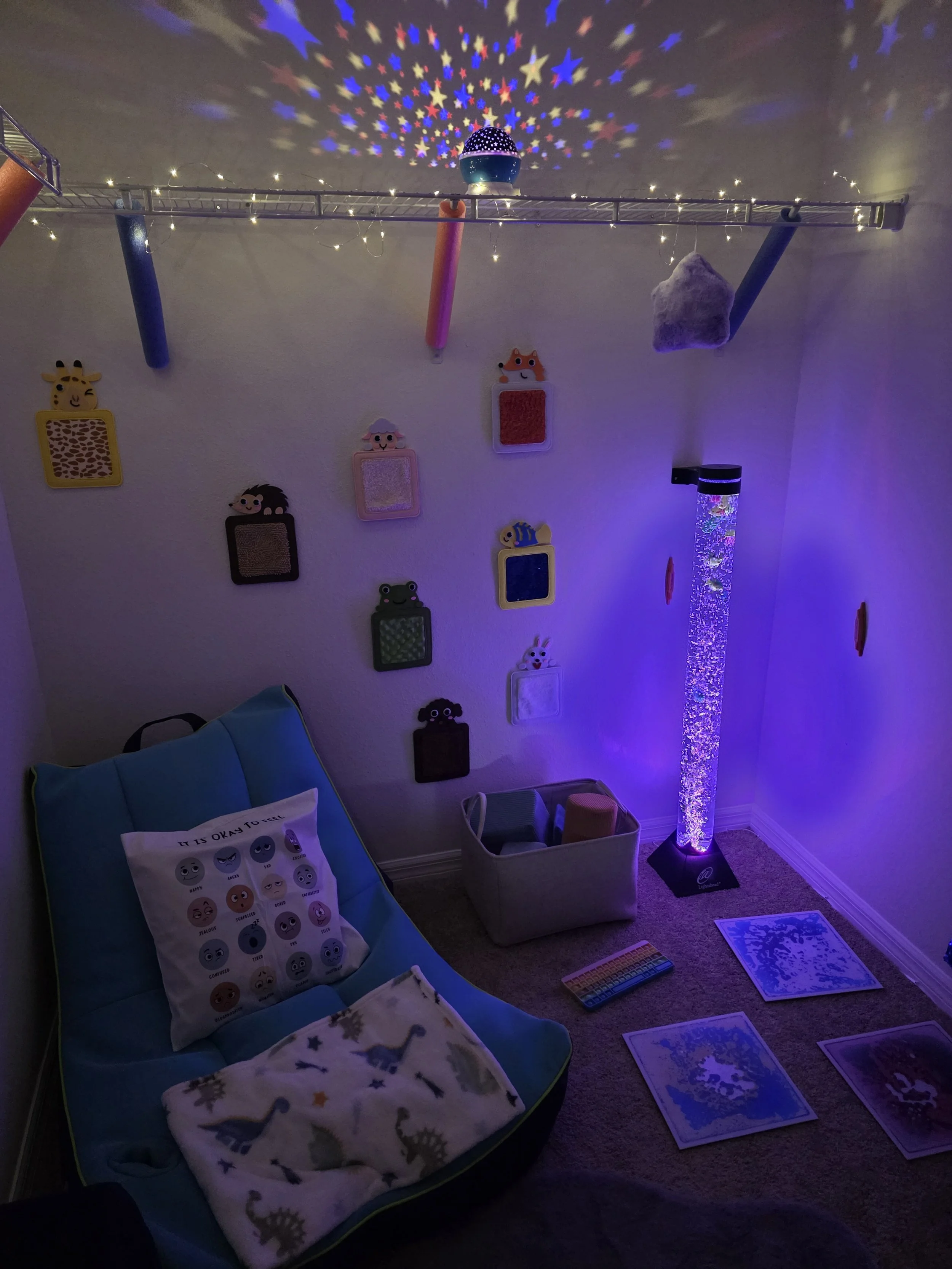 A cozy children's room illuminated with blue and purple lighting, featuring a bubble tube with bubbles and LED lights, a bean bag chair with playful pillows, wall decorations of colorful animal frames, floating foam noodles, and a ceiling projection 