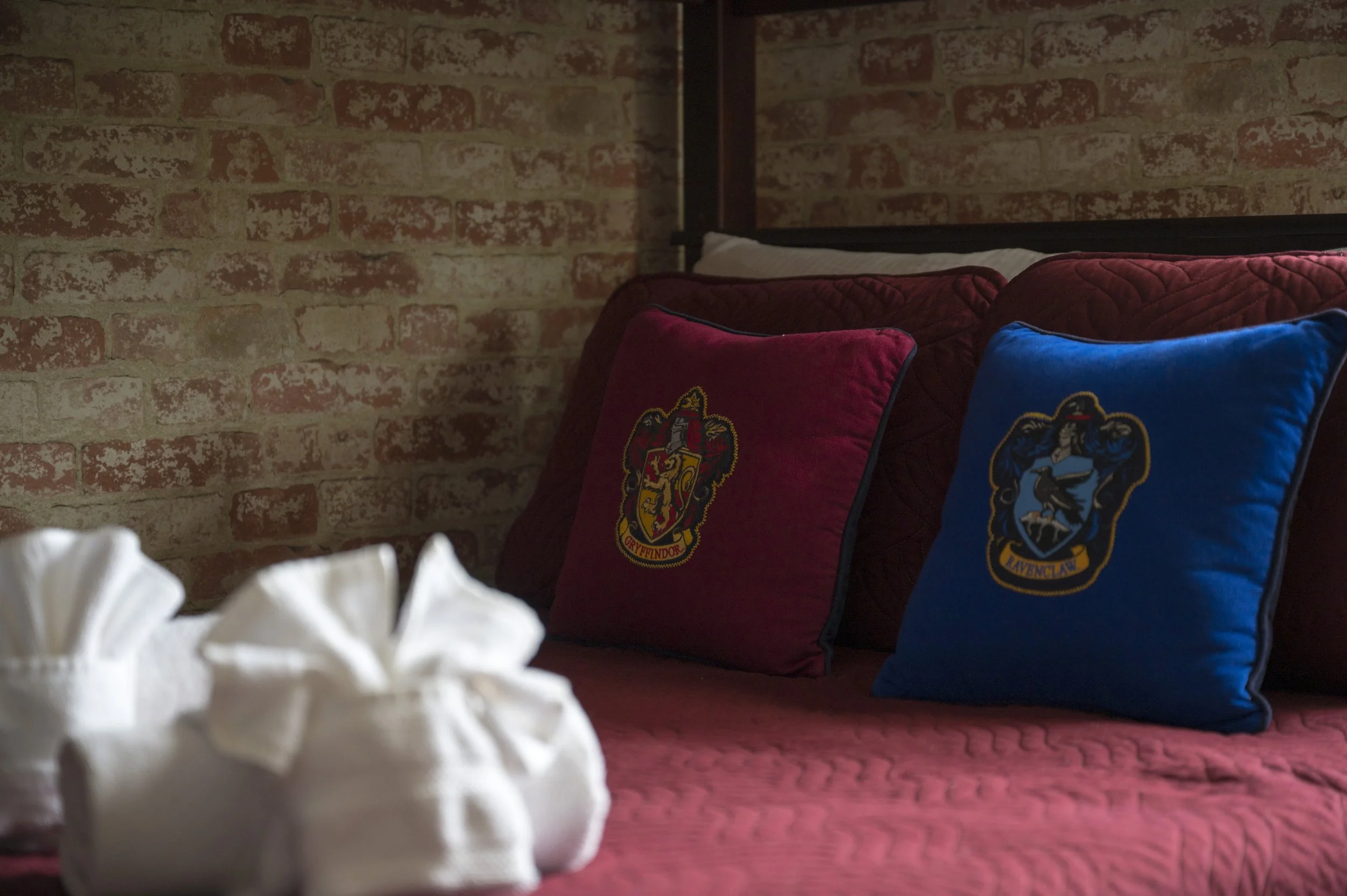 Two pillows with Harry Potter house crests on a bed with a red quilt, against a brick wall, with white towels folded on the side.