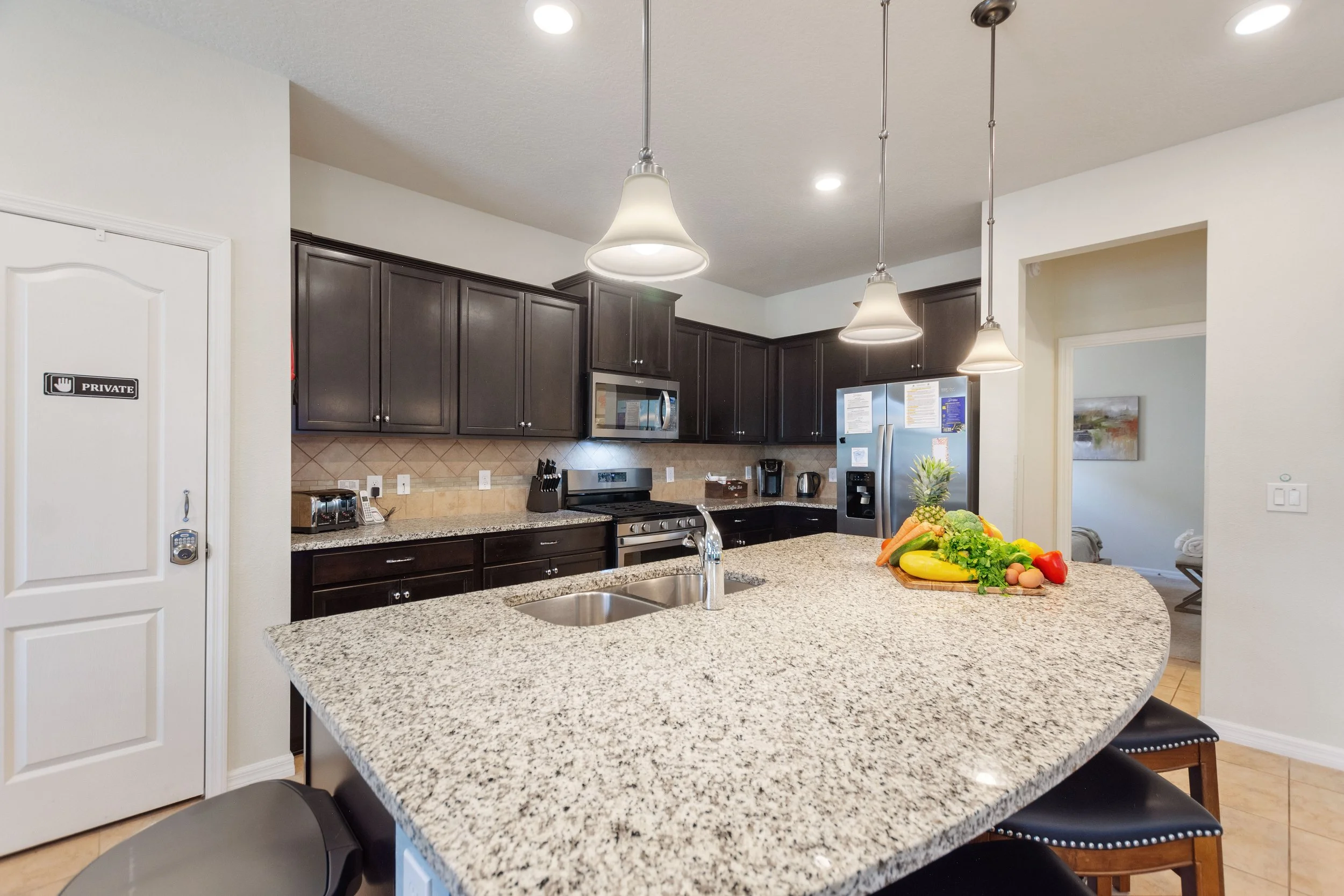 Modern kitchen with dark wooden cabinets, granite countertops, stainless steel appliances, and a large island with a sink. There is a bunch of fresh fruit and vegetables on the island. Pendant lights hang over the island, and a partially open door ha