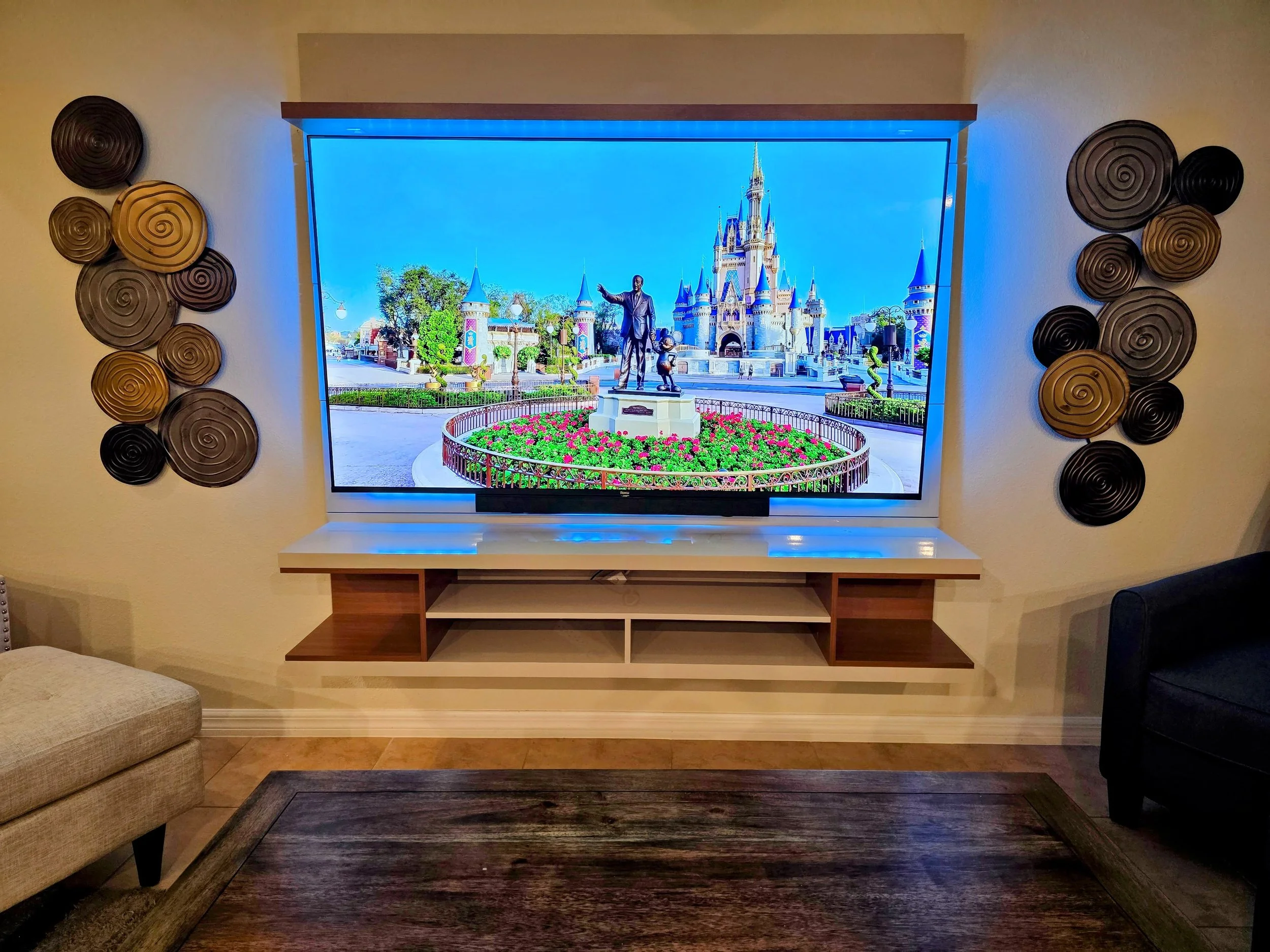 Living room with a wall-mounted digital screen showing a Disney theme park castle and statue, decorated with black and gold spiral wall art on either side, and a beige armchair and black sofa nearby.