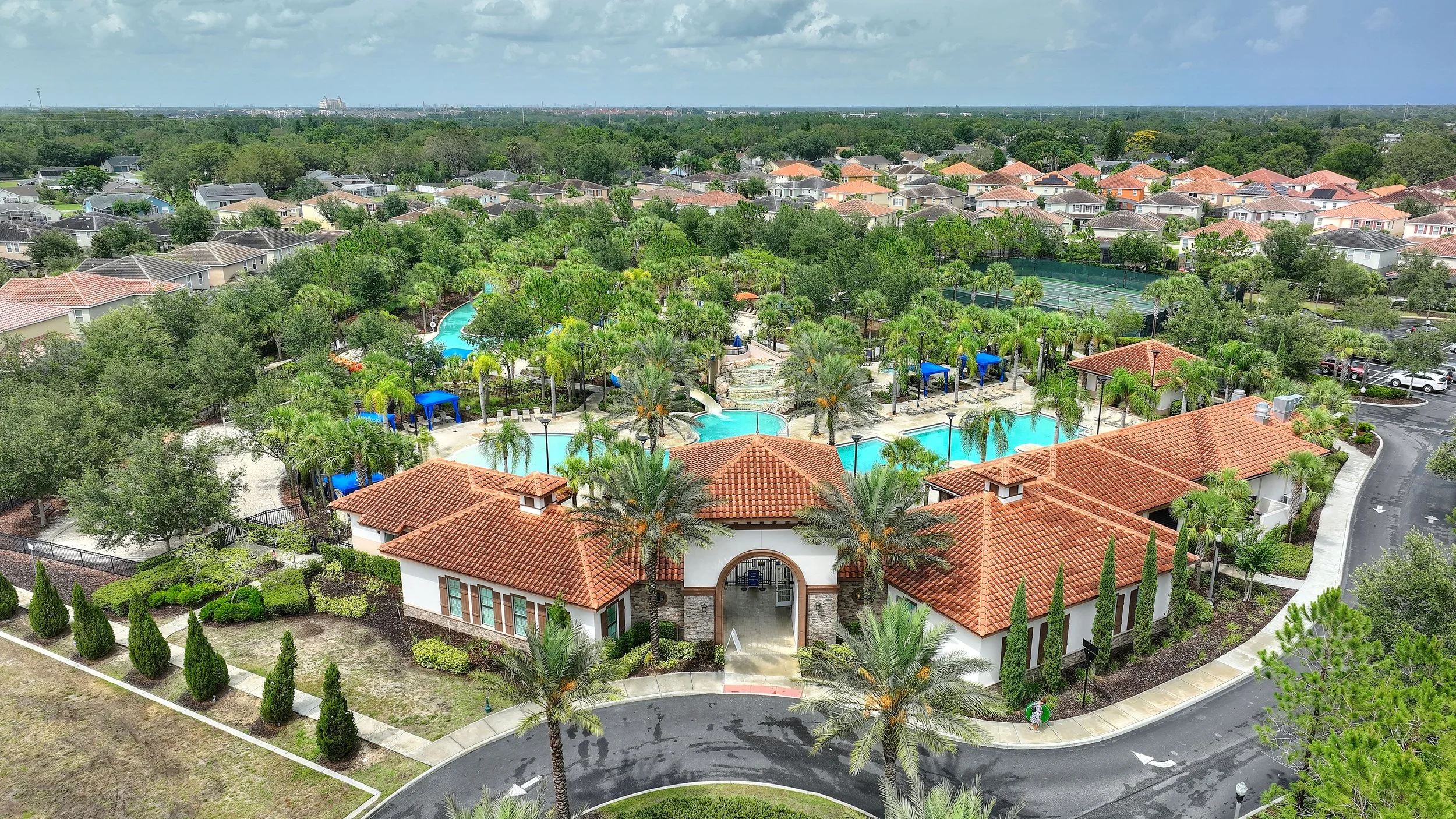 Aerial view of a residential community with a central pool area, surrounded by trees, shrubs, and poolside amenities, with houses in the background.