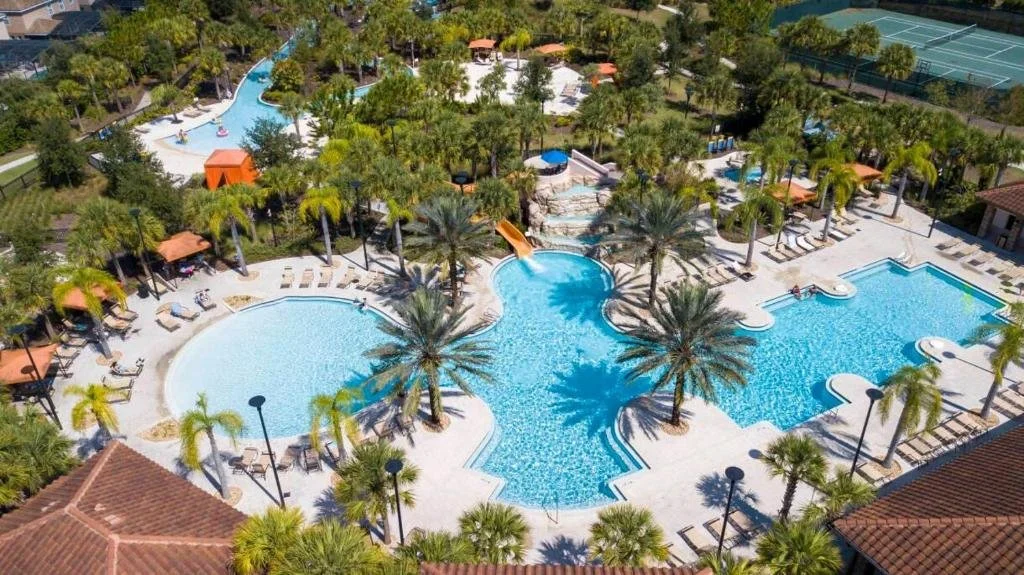 Aerial view of a resort swimming pool area with multiple pools, palm trees, shaded lounging areas, tennis courts, and surrounding buildings.