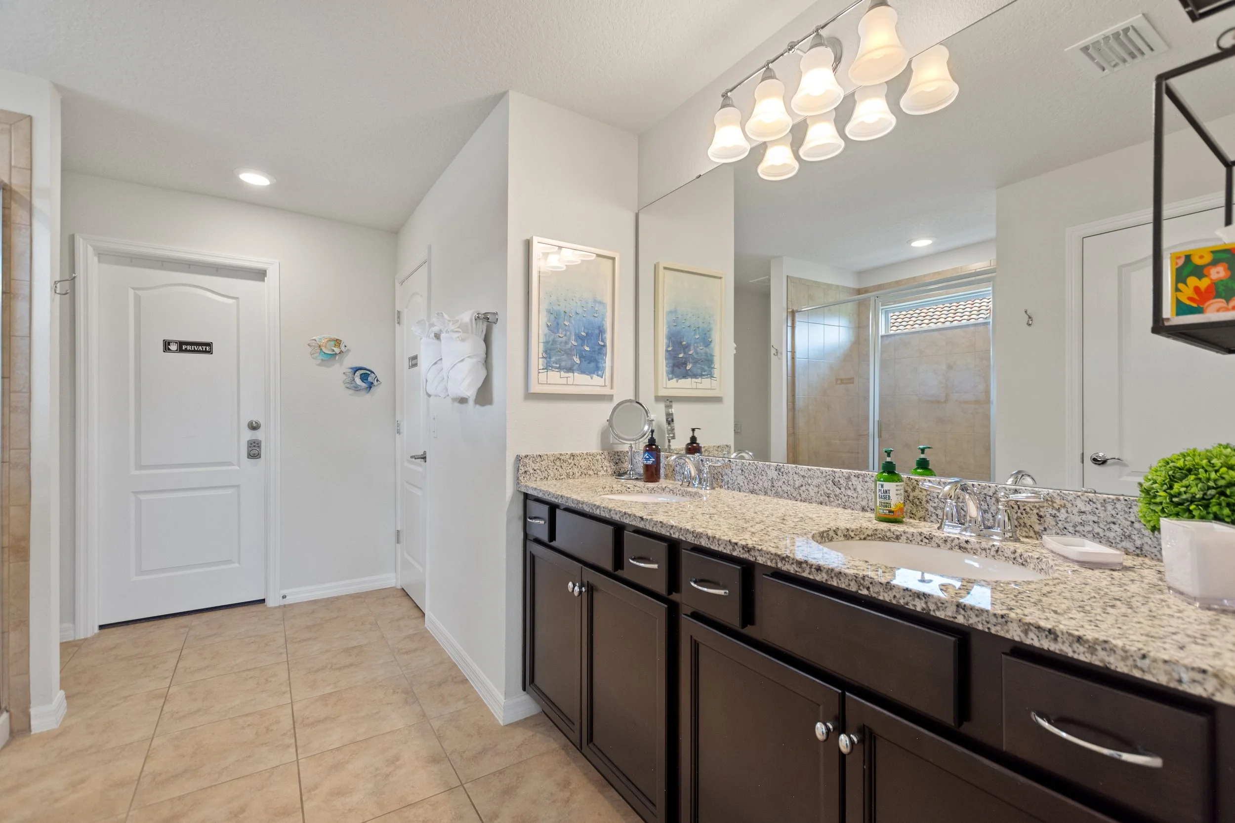 Bathroom with dark wood vanity, granite countertop, dual sinks, large mirror, wall art, towels, soap dispenser, and a separate shower area with a glass door.