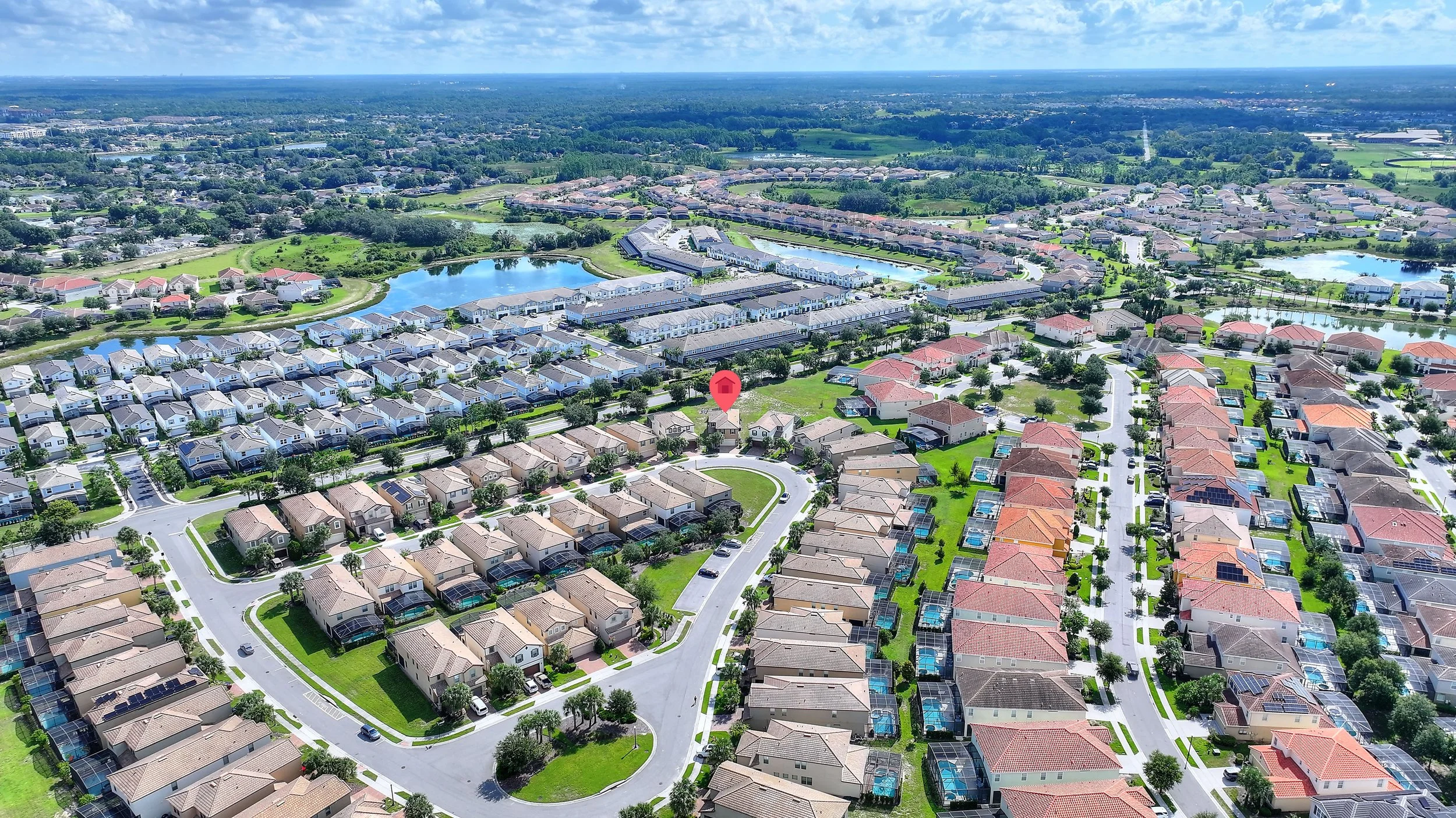 Aerial view of a suburban neighborhood with houses, streets, green lawns, and water features under a partly cloudy sky.