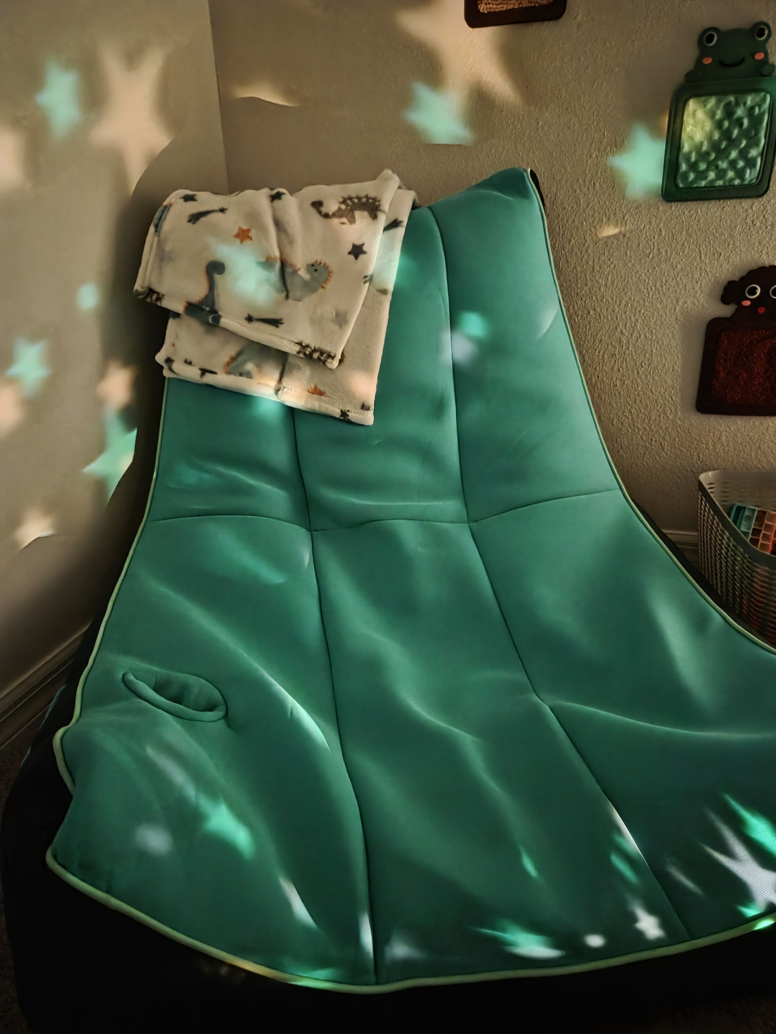 A bright green bean bag chair with a blanket of space-themed dinosaur designs on top, beside a laundry basket filled with items, in a cozy corner with frog and cloud-shaped wall decor, illuminated by star-shaped light reflections.