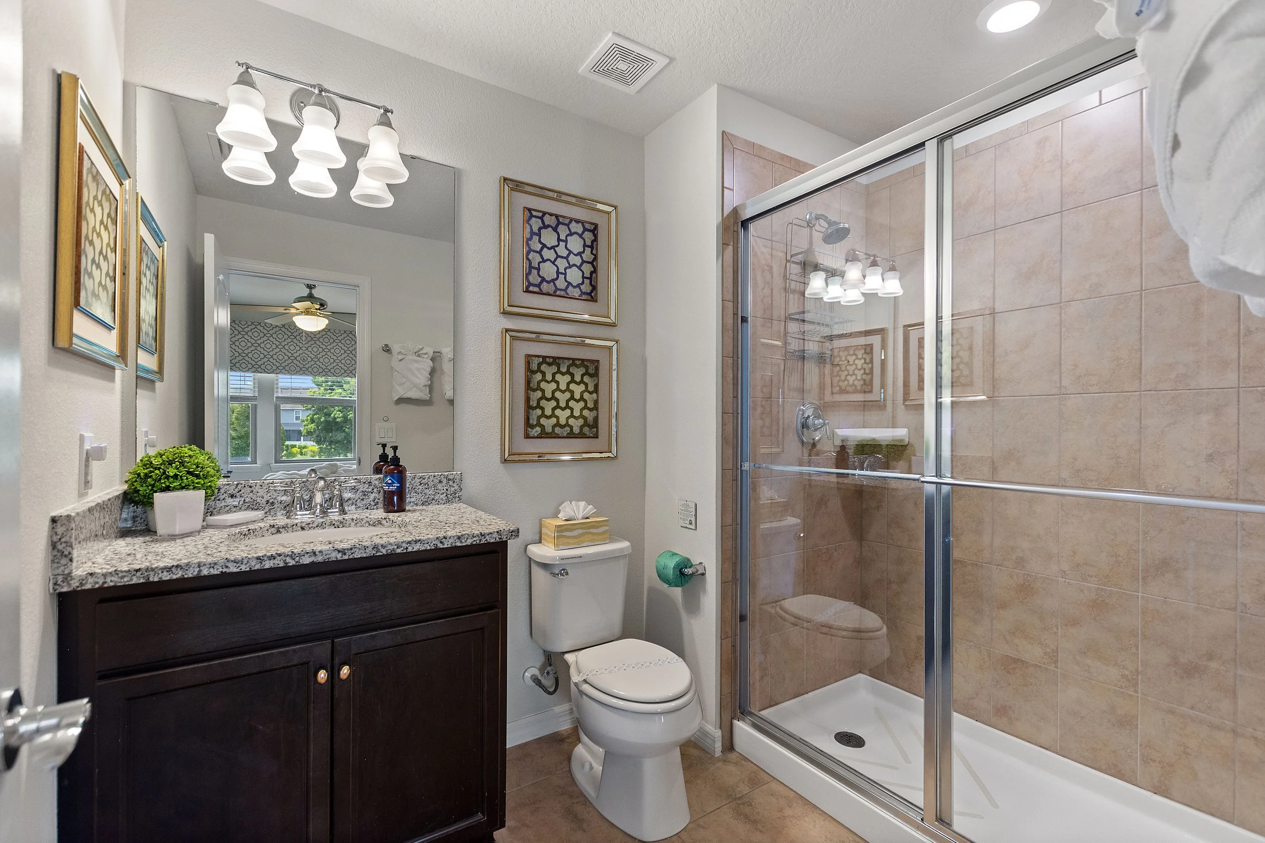 Bathroom with a granite countertop sink, a mirror, a dark wooden vanity, a toilet with tissues on top, framed artwork, a shower with glass doors, tan tiles, and a window with trees outside.
