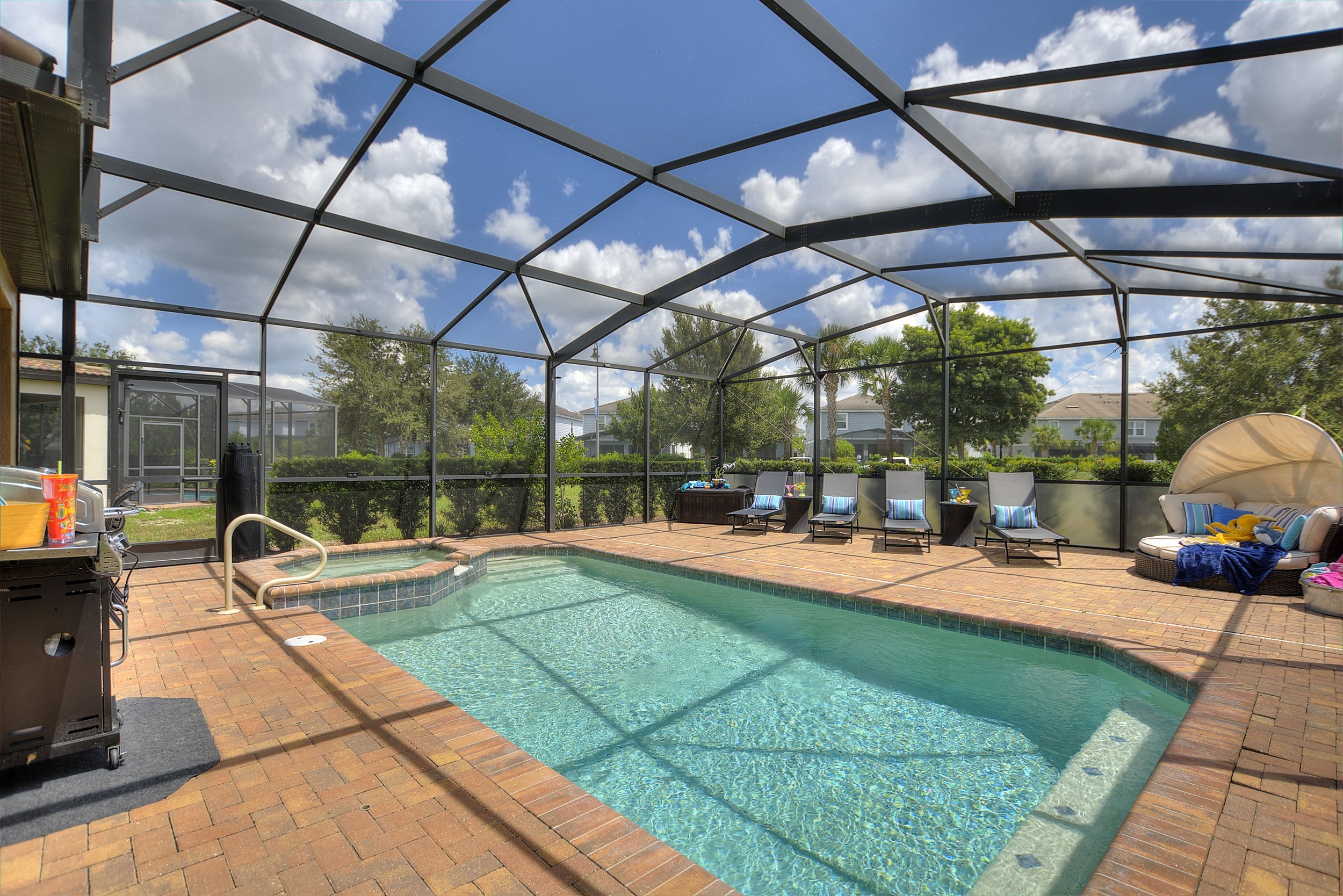 Enclosed backyard swimming pool area with brick decking, pool chairs, a shaded cabana, and a blue sky with scattered clouds.
