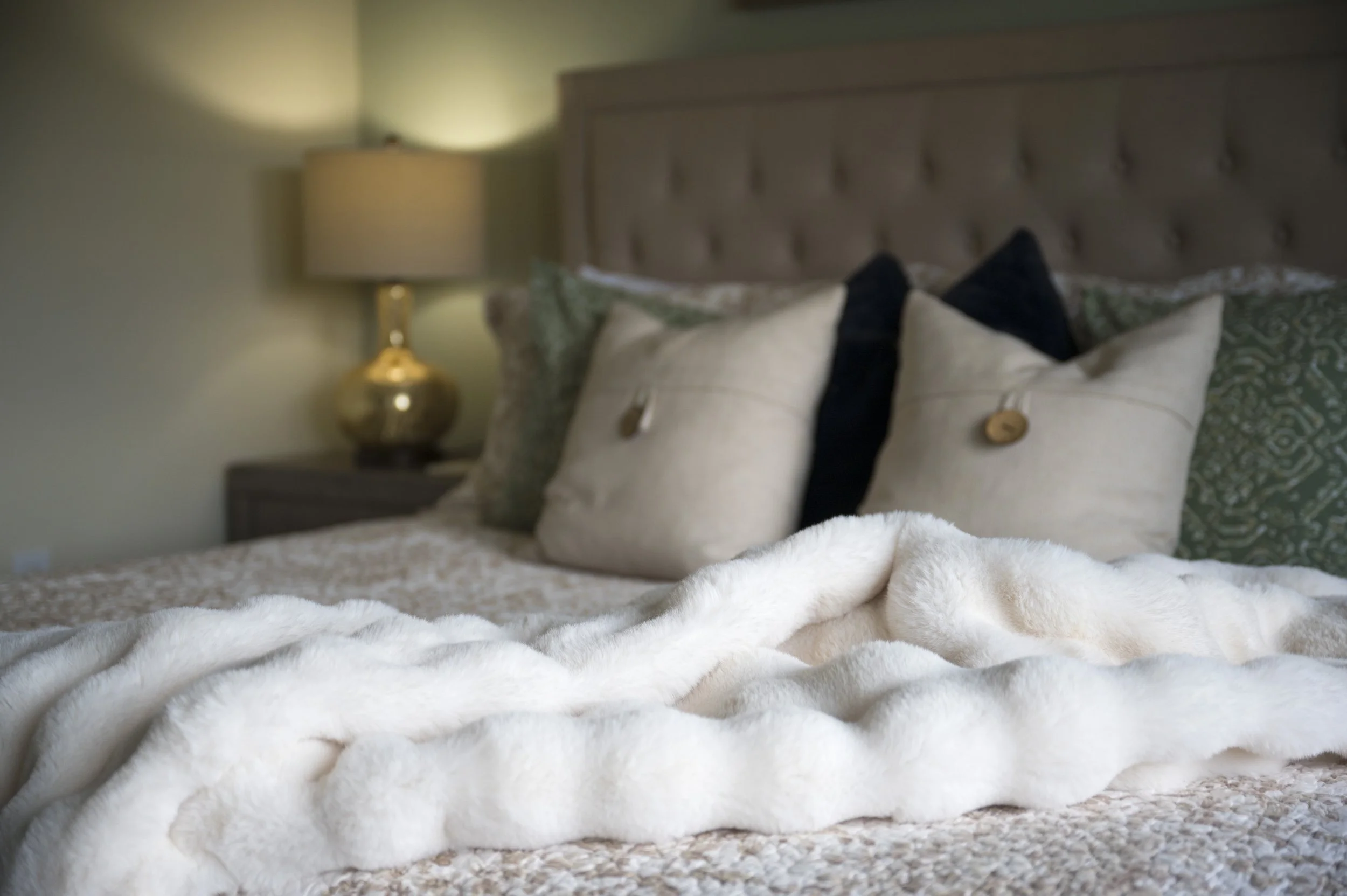 A neatly made bed with decorative pillows and a beige blanket in a bedroom with a nightstand and a lamp.