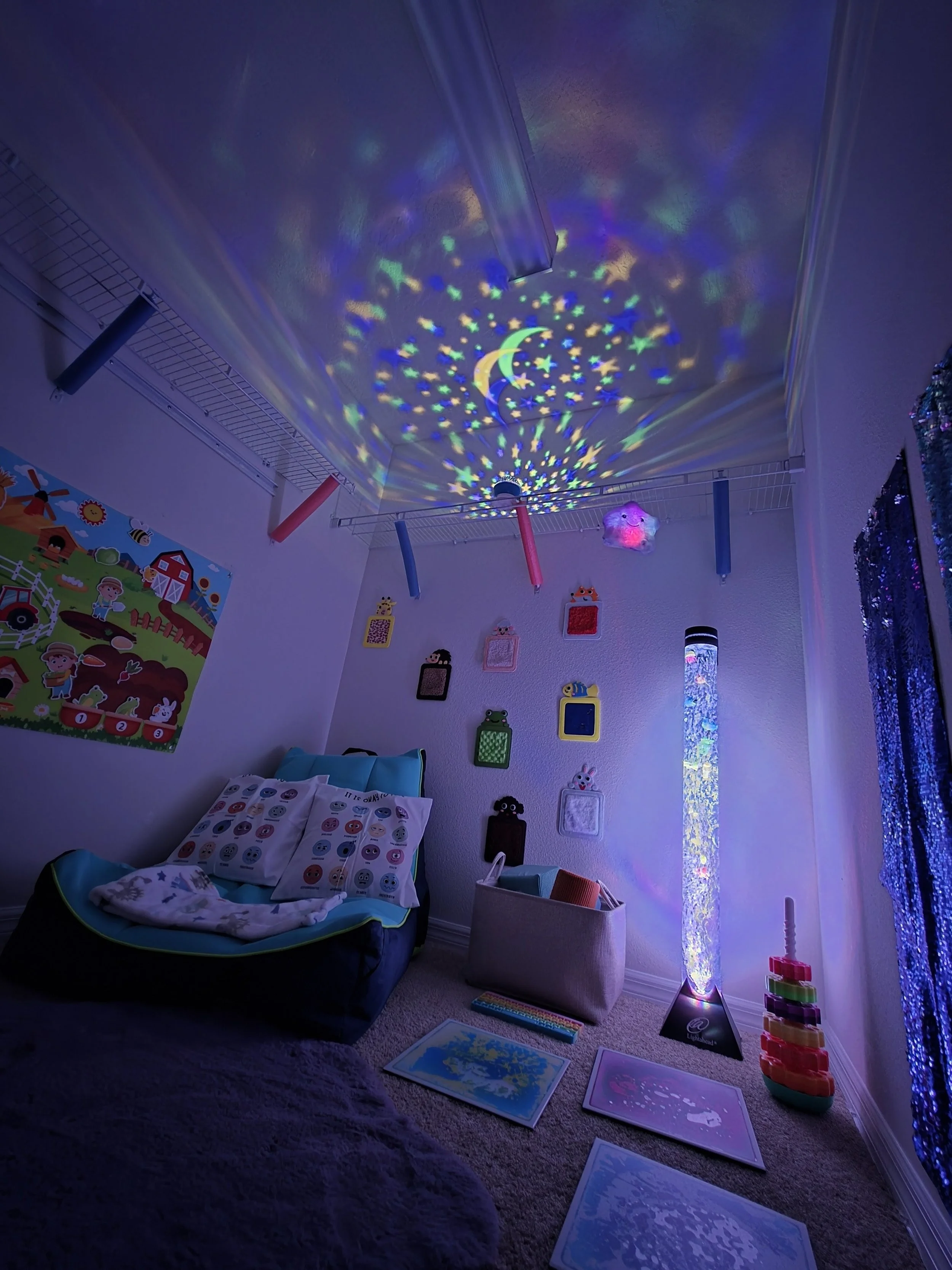 A child's decorated bedroom with a star and moon projector casting colorful light patterns on the ceiling, artwork on the wall, and a cozy seating area with pillows and a blanket.