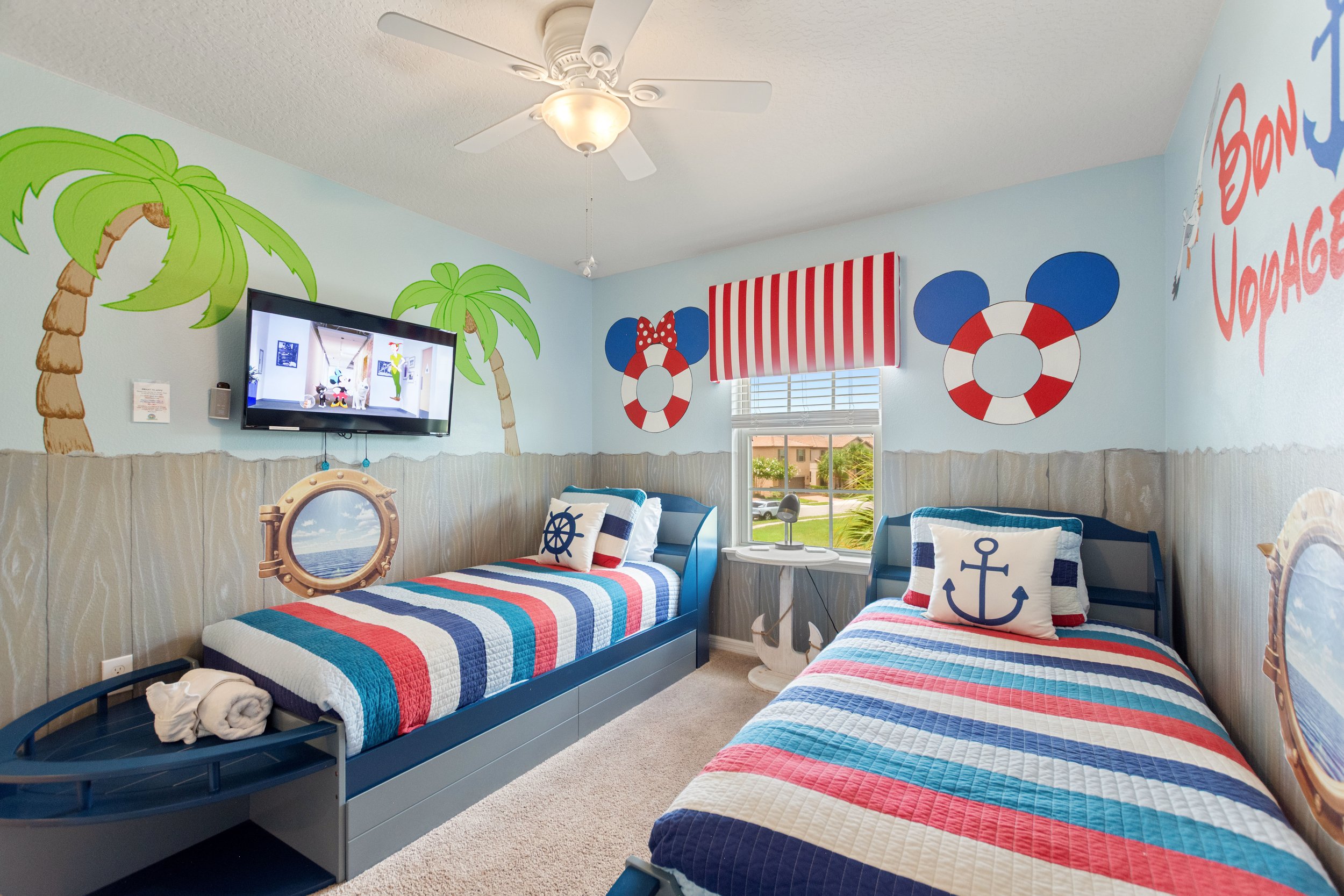 A children's bedroom with beach and nautical theme decor. It features two twin beds with striped quilts in red, white, and blue, nautical pillows, a window with a striped valance, and wall art including palm trees, life preservers, and the phrase 'Bo