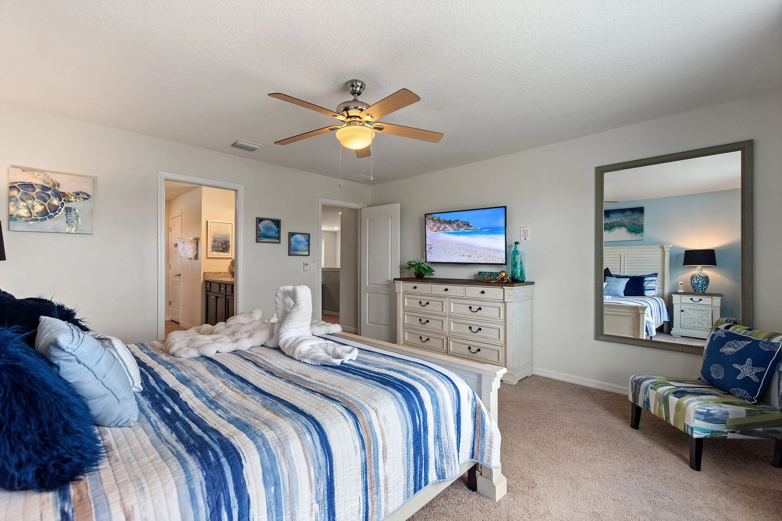 A bedroom with a large bed covered in blue striped bedding and decorative pillows, a mirror reflecting another bed, a dresser with a television, and ocean-themed decor.