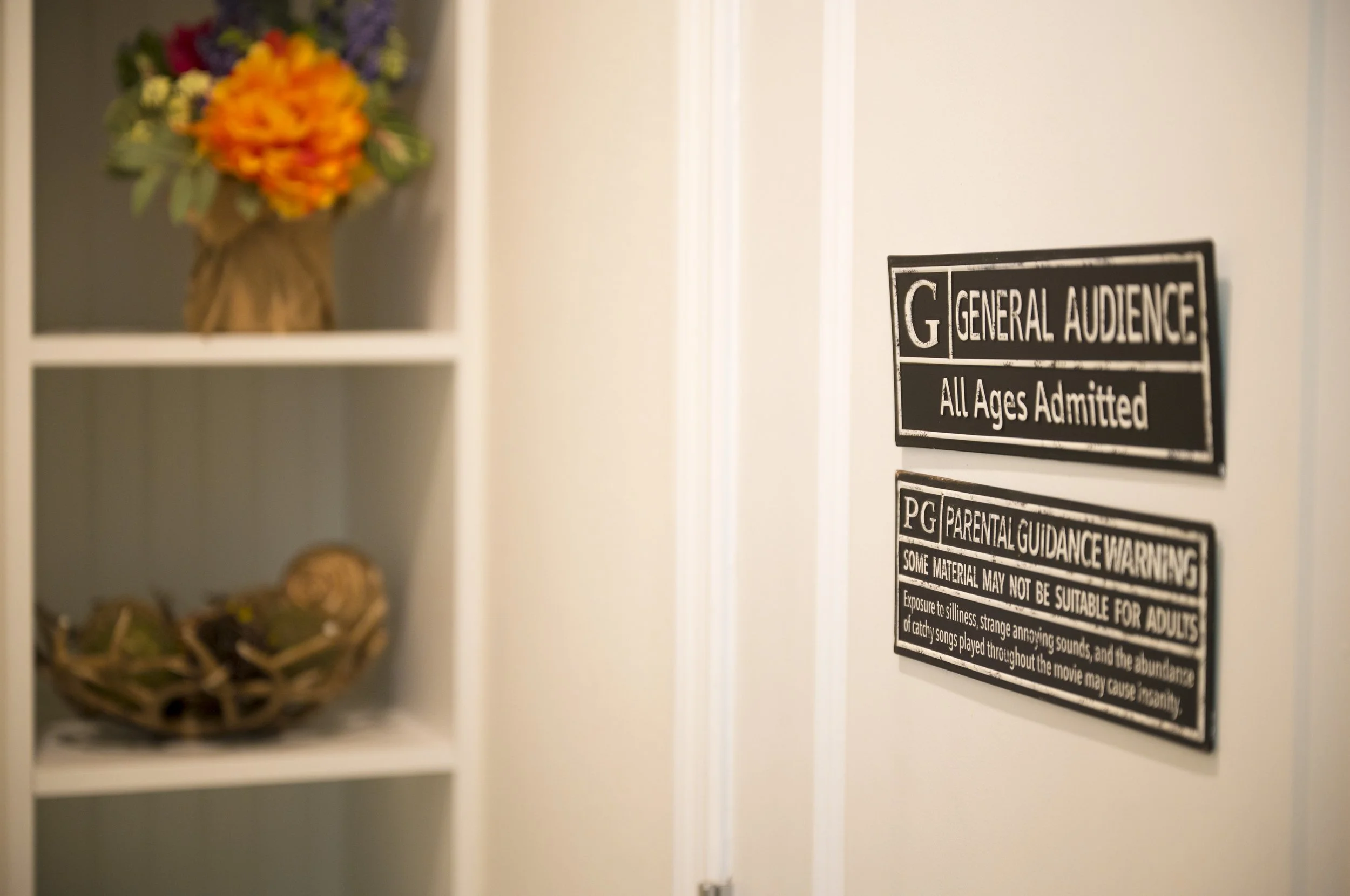 Close-up of a movie theater door with age restriction signs and a bookshelf with a flower arrangement in the background.