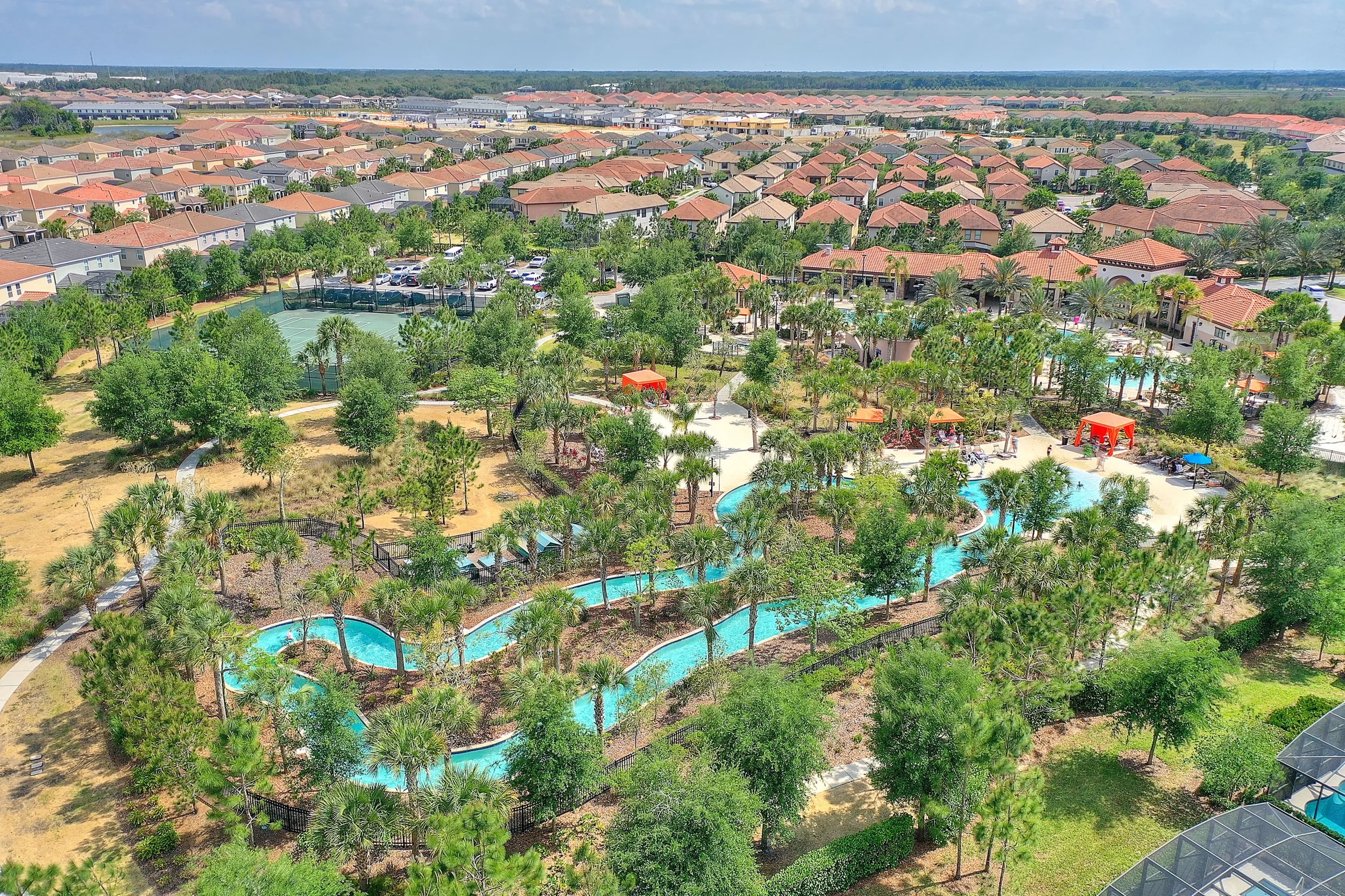 Aerial view of a community with houses, a swimming pool, water slide, and tennis courts, surrounded by trees and landscaping.
