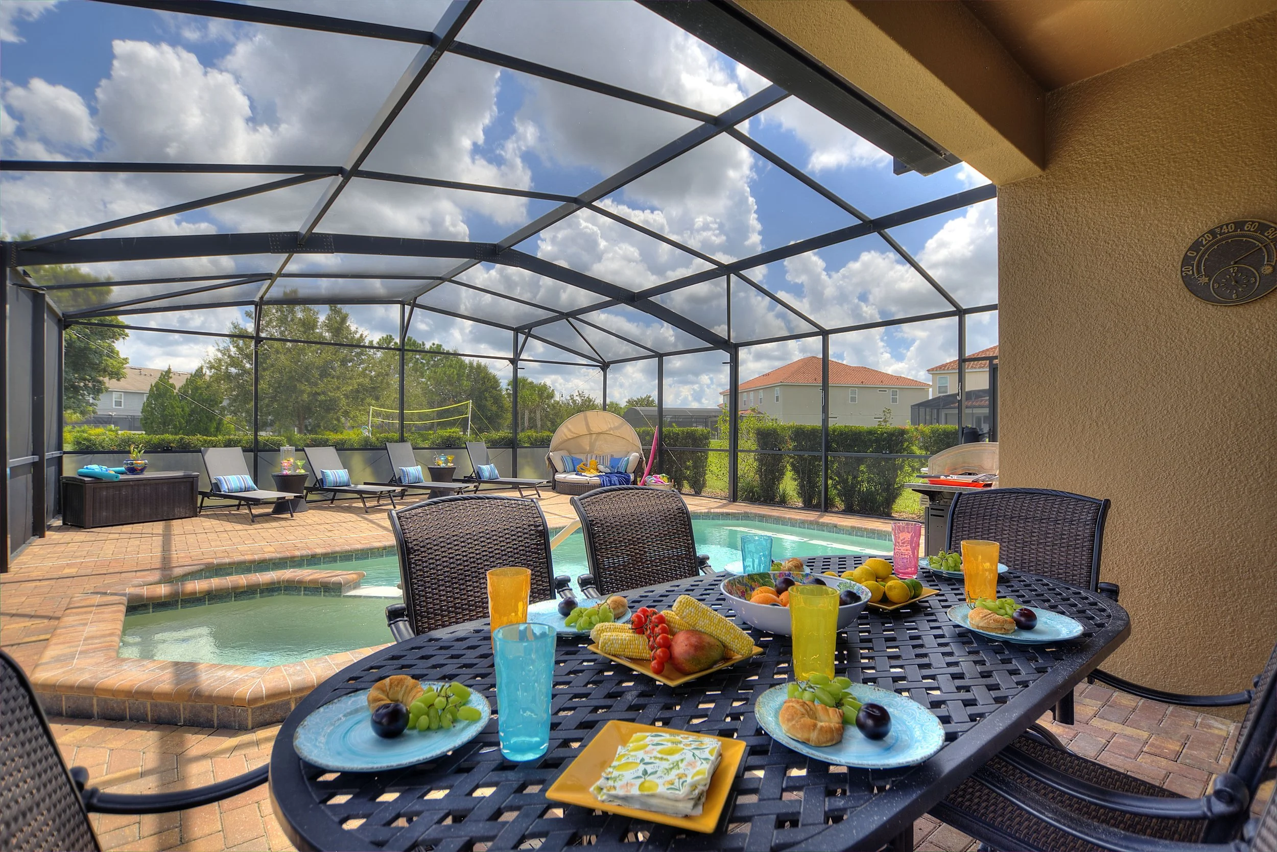 Outdoor screened-in patio with a swimming pool, sun loungers, a dining table set with food and drinks, and a cloudy sky.