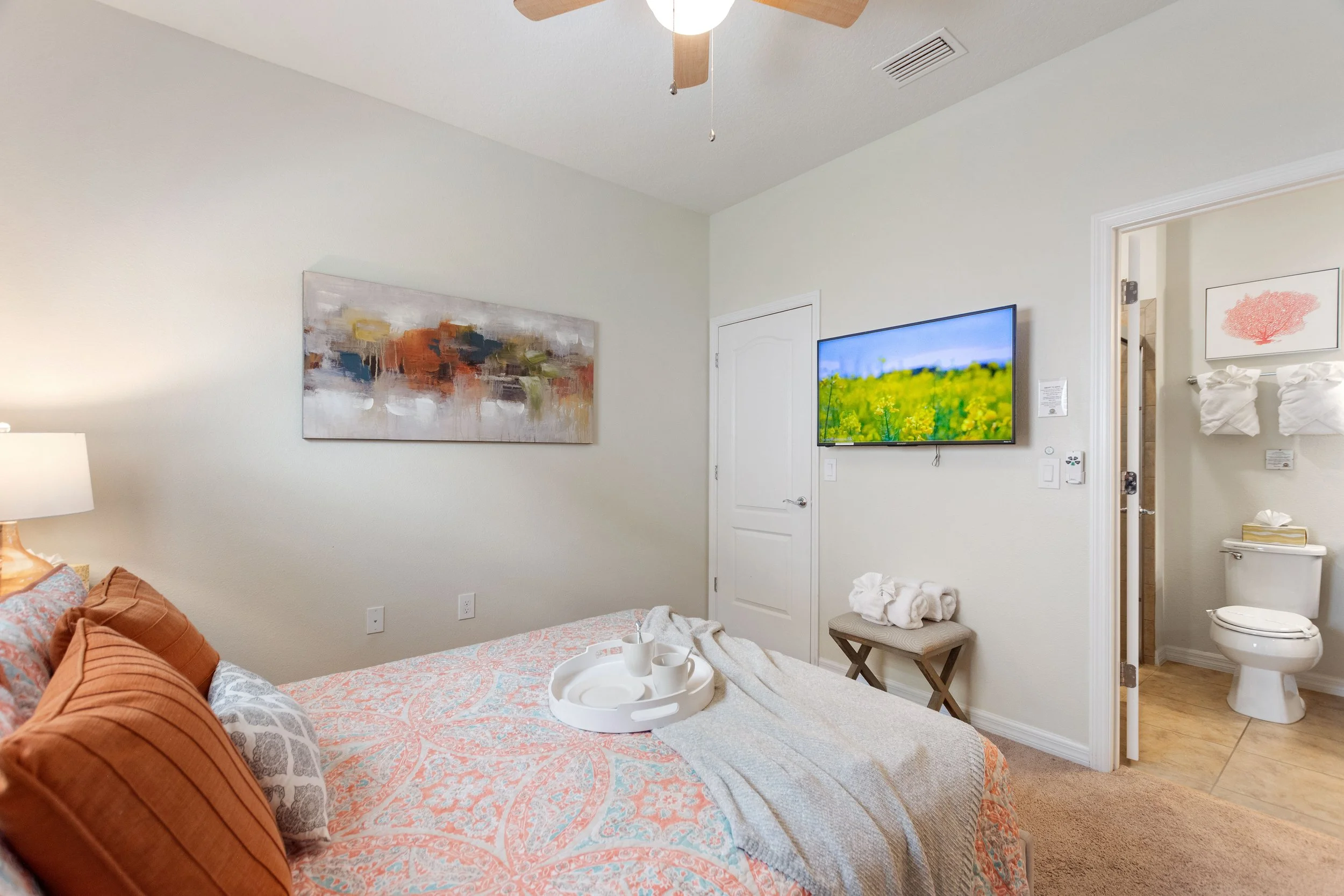 A bedroom with a bed covered in a patterned bedspread and pillows, a ceiling fan, a wall-mounted TV displaying a nature scene, abstract wall art, a nightstand with a lamp, towel art on the bathroom wall, a toilet, and an open door leading to the bath