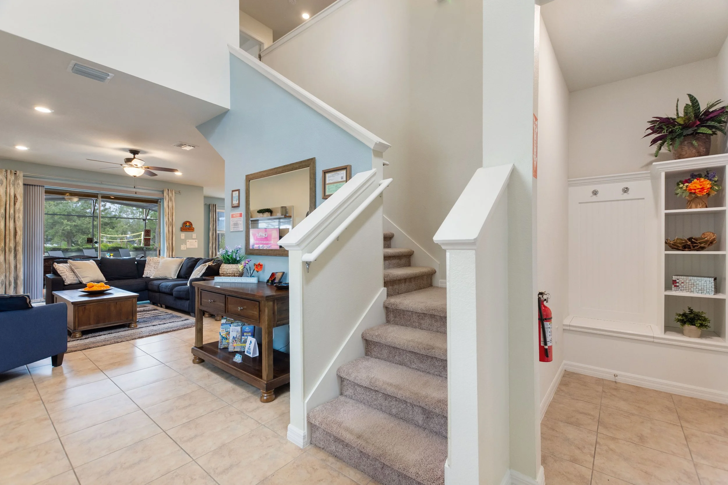Living room with tiled floor, blue and beige furniture, beige curtains, wood coffee table, window view of greenery, staircase with carpeted steps, hallway with built-in shelves, fire extinguisher on wall.
