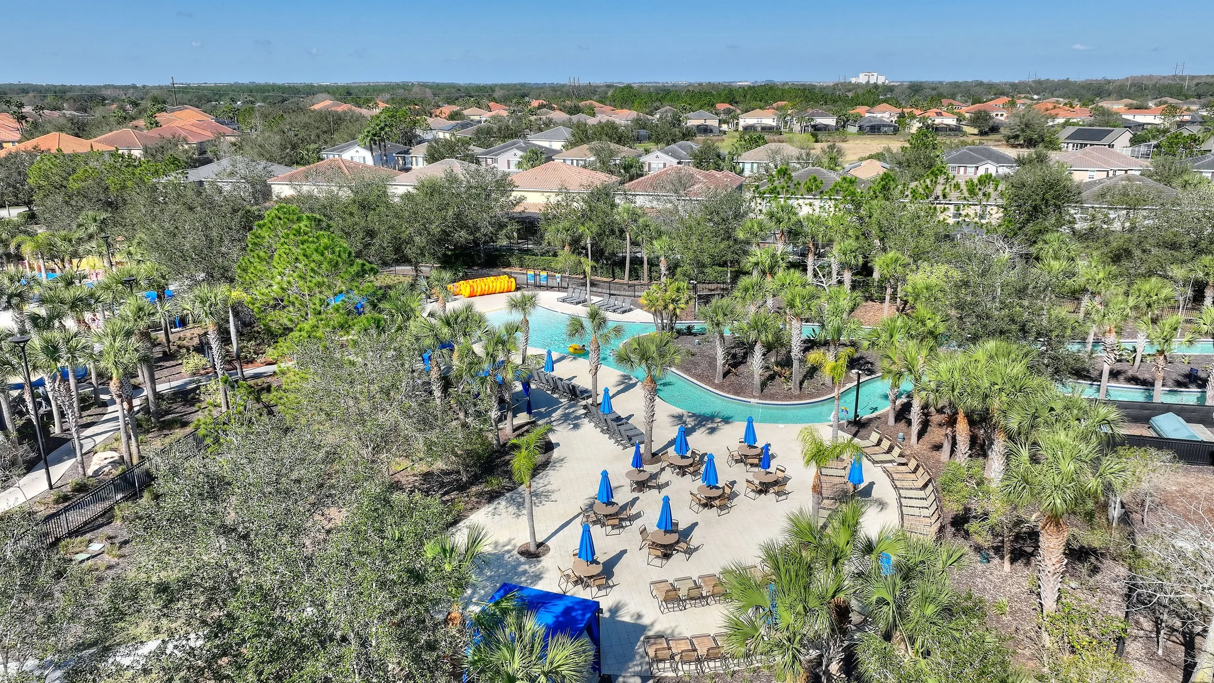 Aerial view of a resort swimming pool area surrounded by palm trees and lounge chairs with blue umbrellas, residential houses in the background.