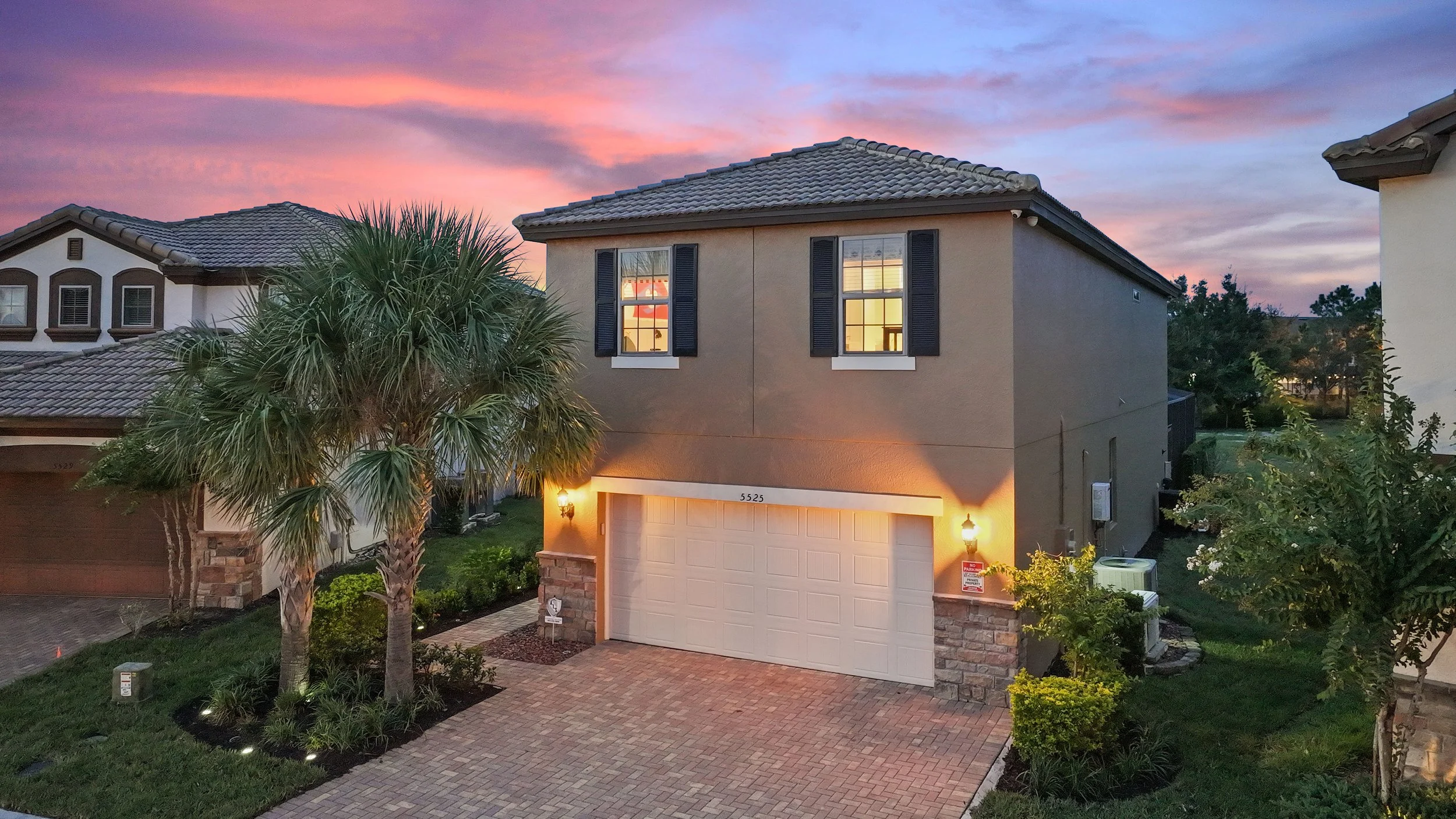 A two-story suburban house at sunset with exterior lights on, a brick driveway, palm trees in the front yard, and a sunset sky with pink and purple clouds.