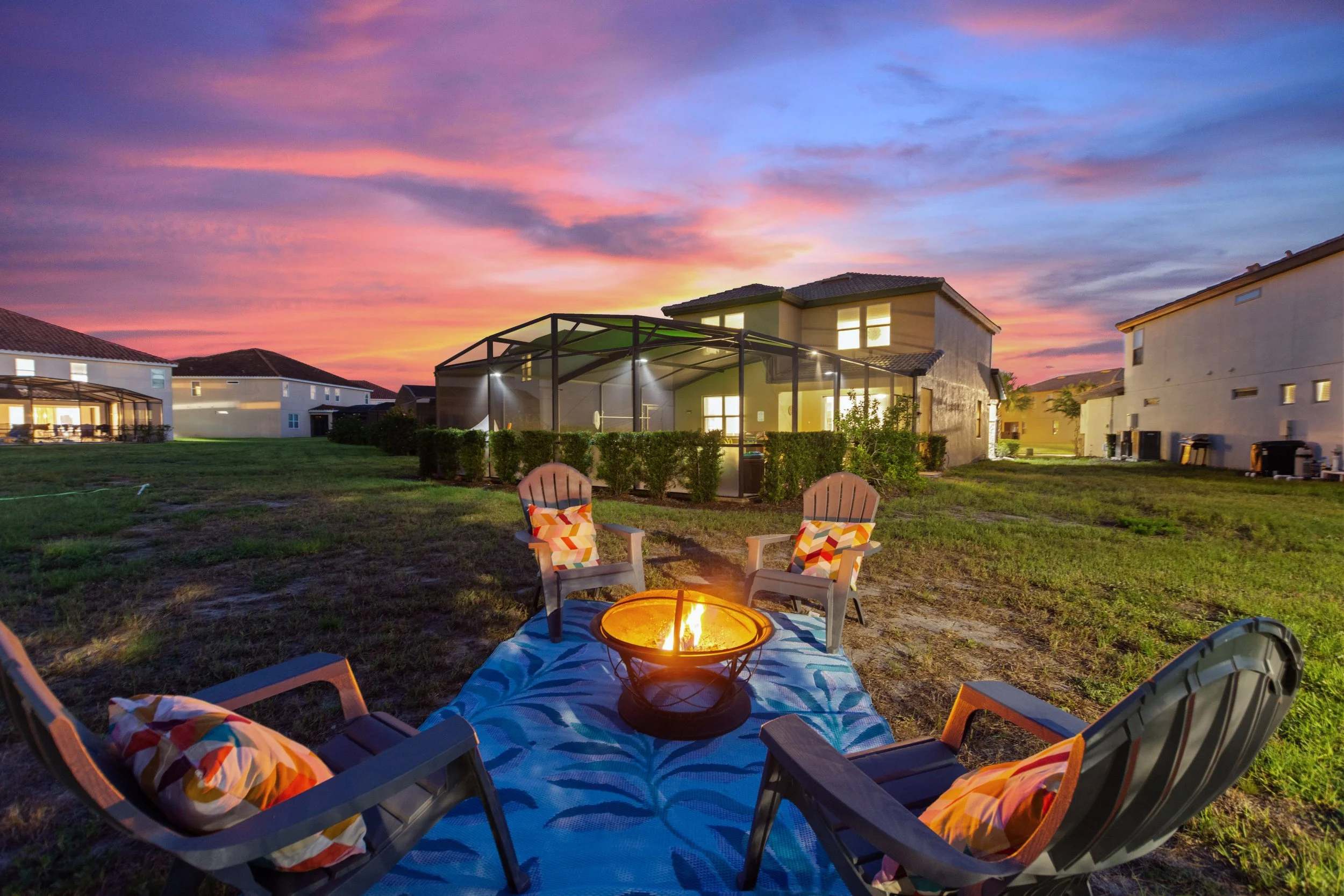 A backyard at sunset with a fire pit surrounded by four chairs with colorful cushions, blue patterned outdoor rug, and neighboring houses in the background.