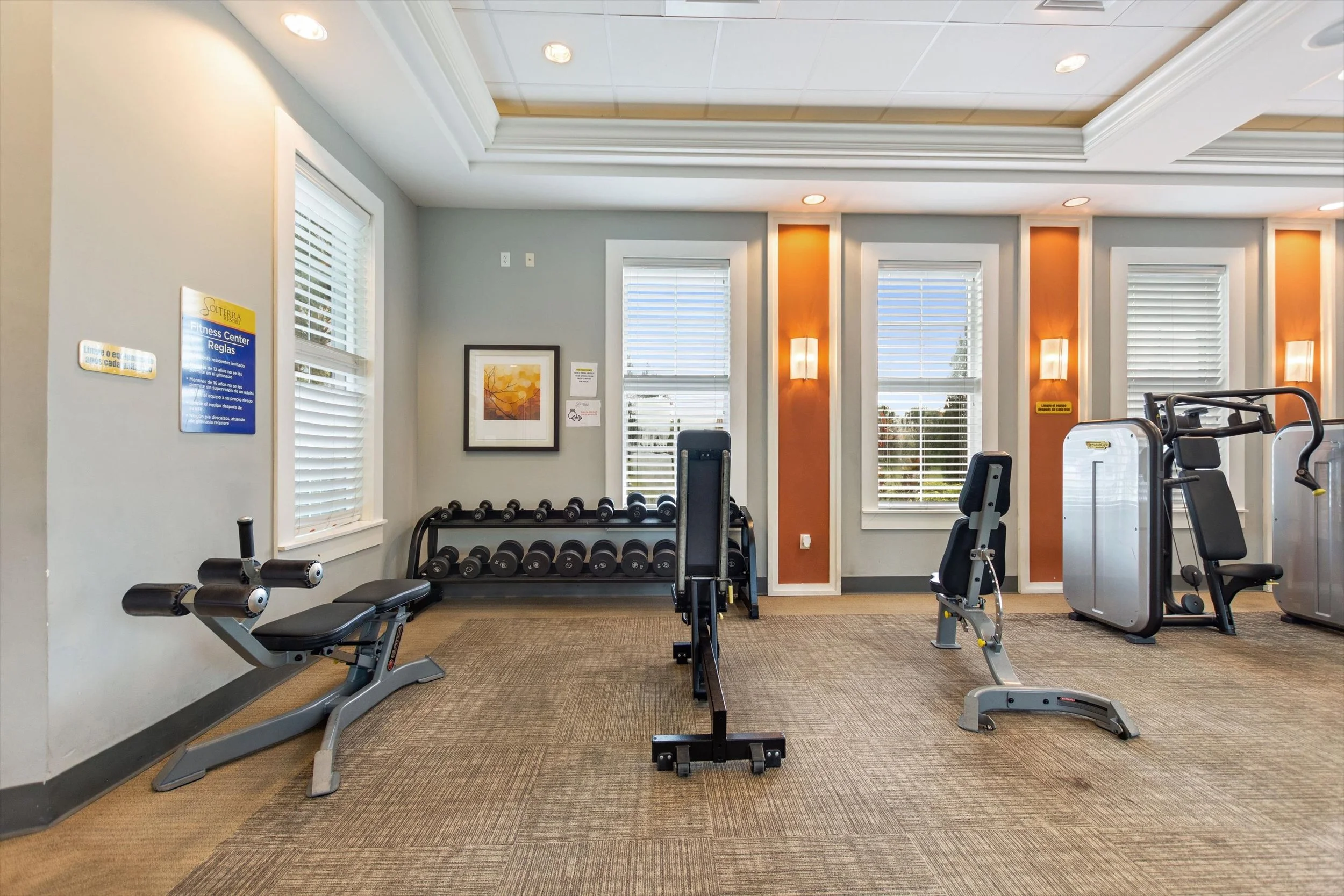 Fitness center with exercise equipment, including a bench, dumbbells on a rack, and workout machines, in a brightly lit room.