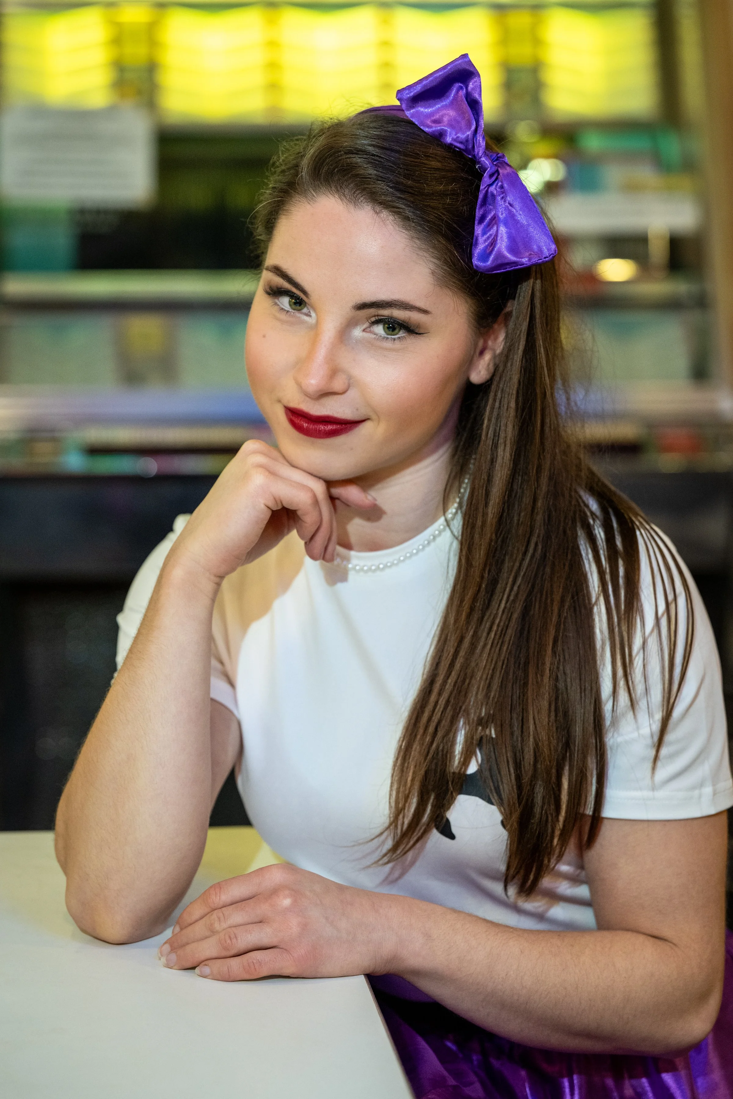 Young woman with long brown hair, wearing a purple bow headband and red lipstick, sitting at a table in a casual setting, with a background of stained glass windows.