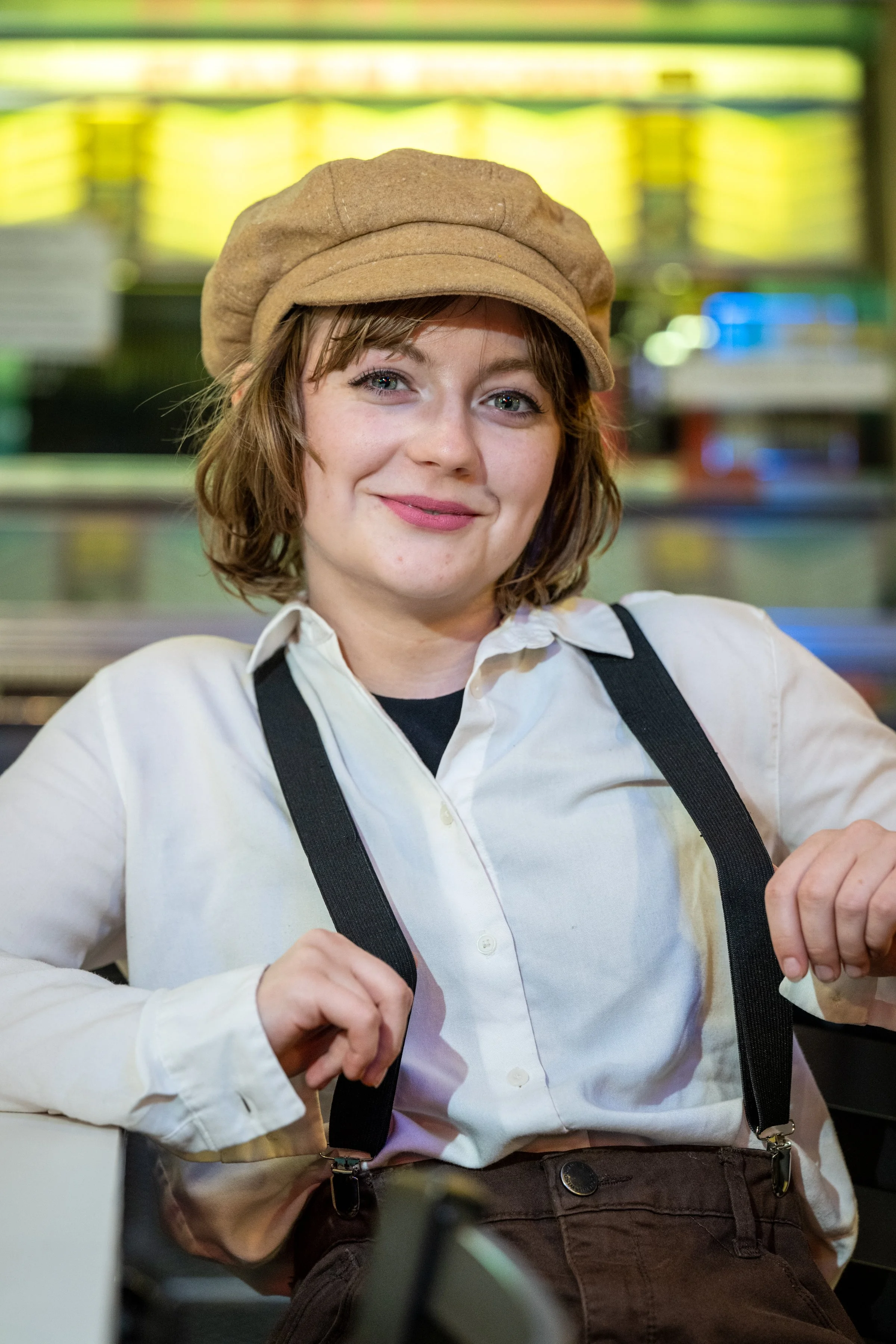 A young woman with short brown hair, wearing a beige beret, white blouse, and suspenders, smiling confidently.