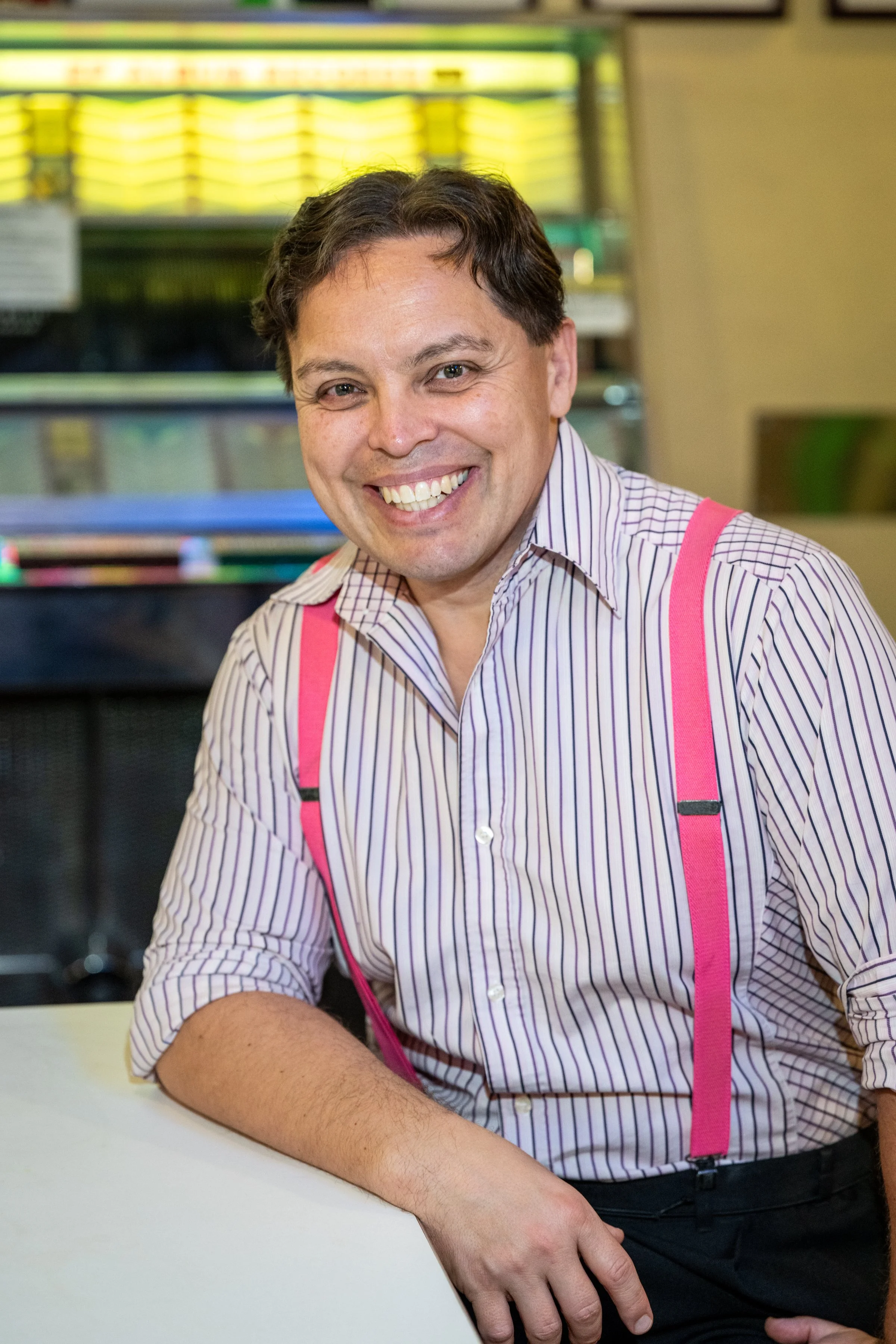A man smiling and sitting at a table in a brightly lit environment, wearing a striped shirt and pink suspenders.