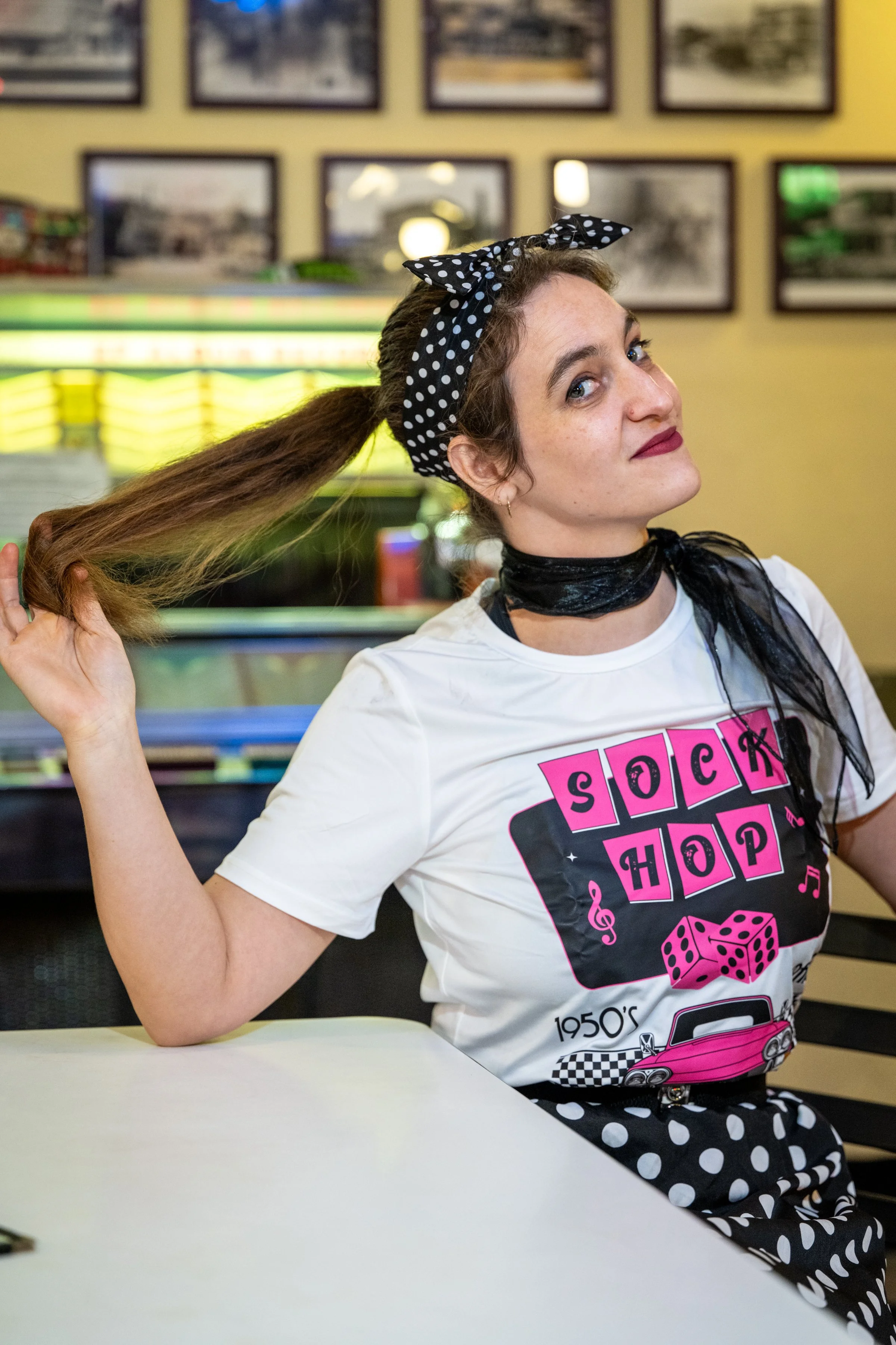 A young woman with dark hair, blue eyes, and dark red lipstick, wearing a white T-shirt with pink and black graphics, a black scarf, and a black and white polka-dotted skirt. She is sitting in a restaurant, holding her long hair with one hand, and looking at the camera with a playful expression.