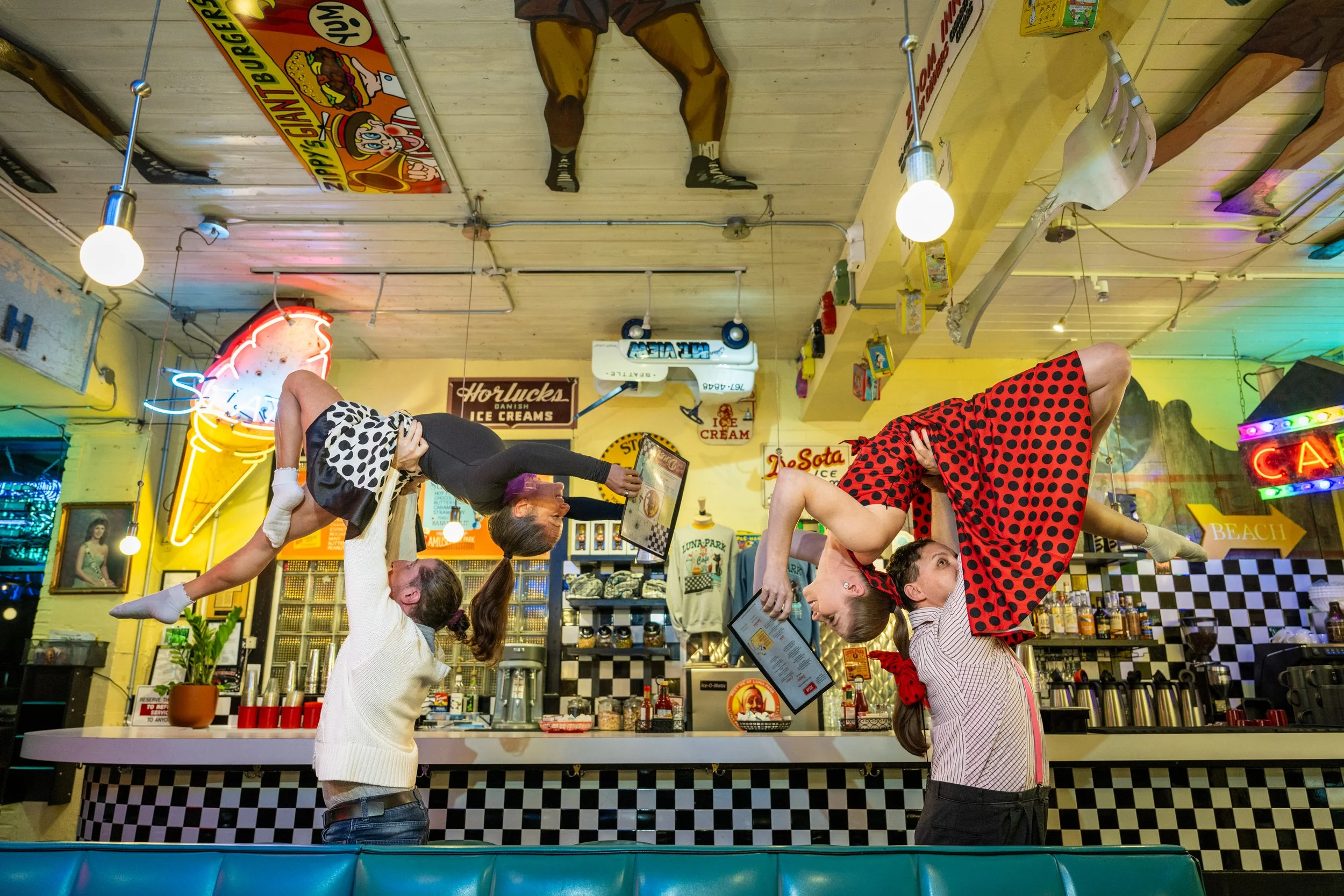 Performers representing diner patrons perform acrobatic lifts. The two women being lifted read the menu while lifted above the heads of the male acrobats.