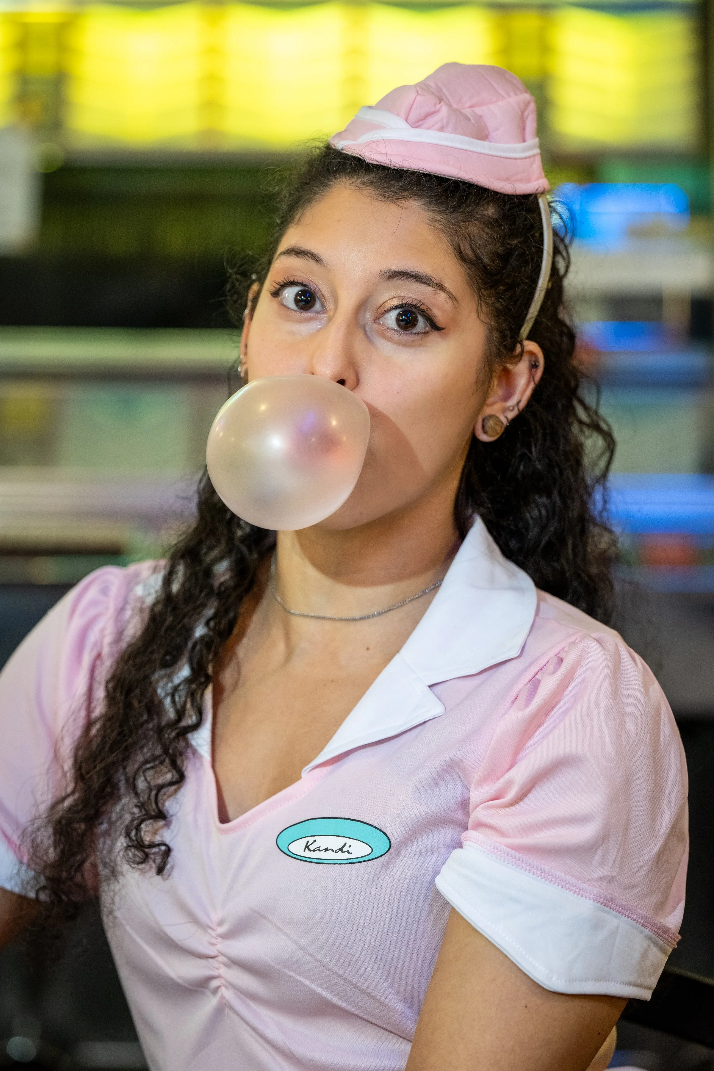 A young woman in a pink waitress uniform blowing a bubblegum bubble, wearing a pink cap and a name tag that says 'Kandi', in an indoor environment with colorful background lights.
