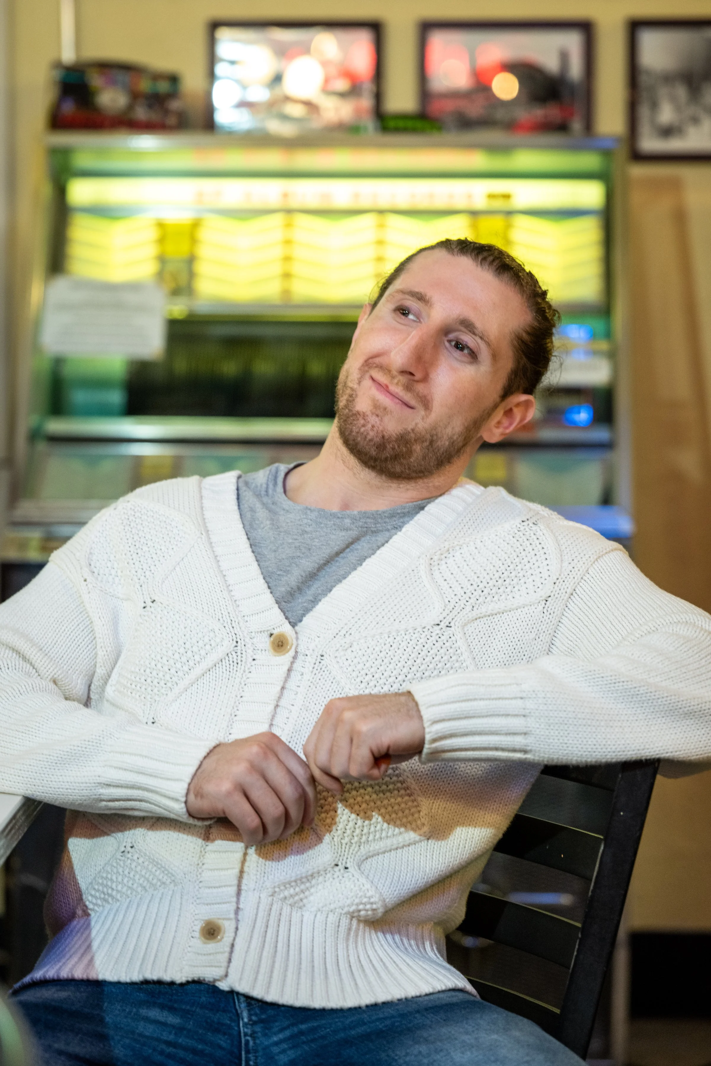 A man with a beard and long hair tied back, sitting in a chair, wearing a white knit cardigan over a gray T-shirt, in what appears to be a diner or cafe with a jukebox in the background.