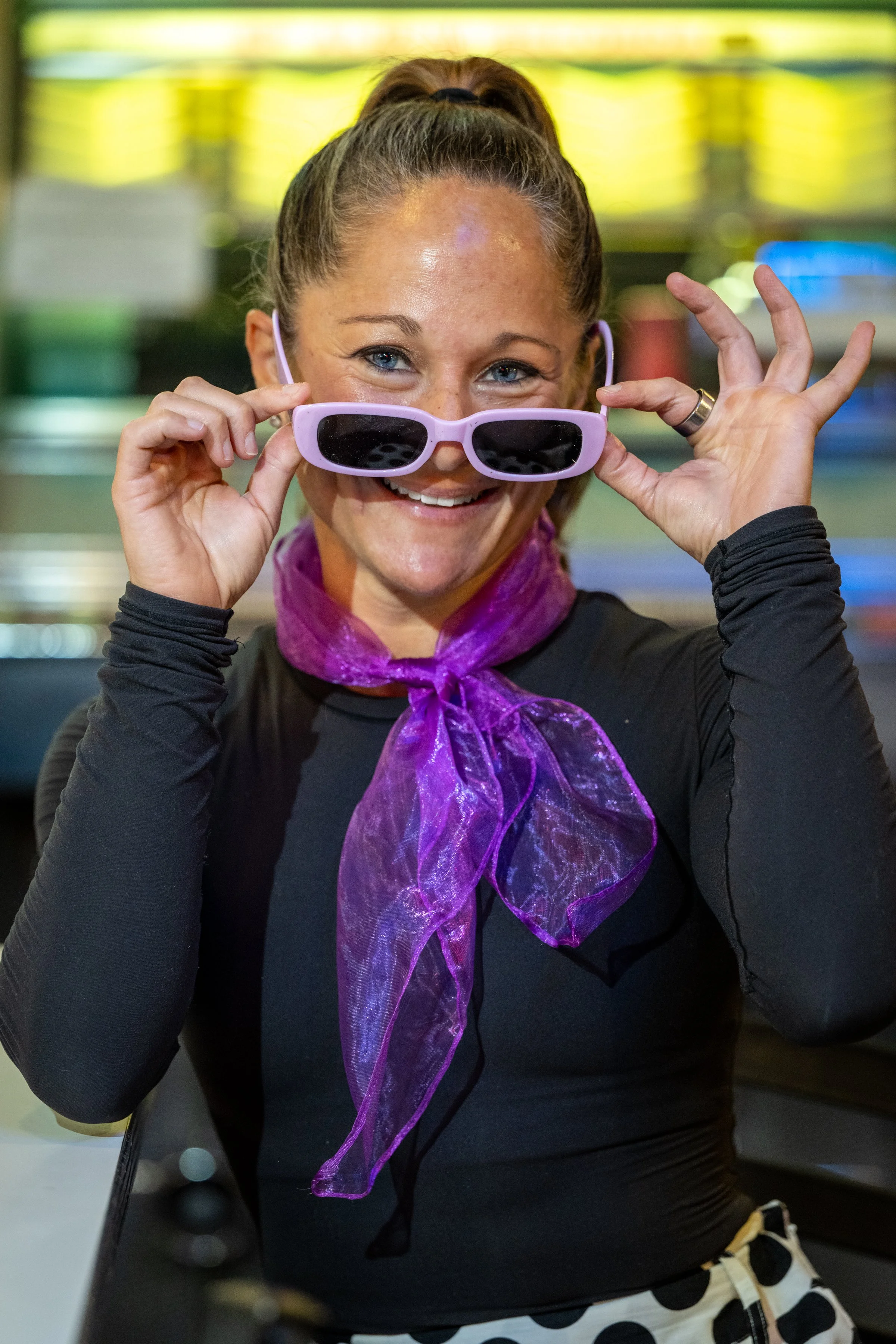 Woman with a high ponytail and purple sunglasses, smiling and holding sunglasses, wearing a black top and purple scarf, indoors.