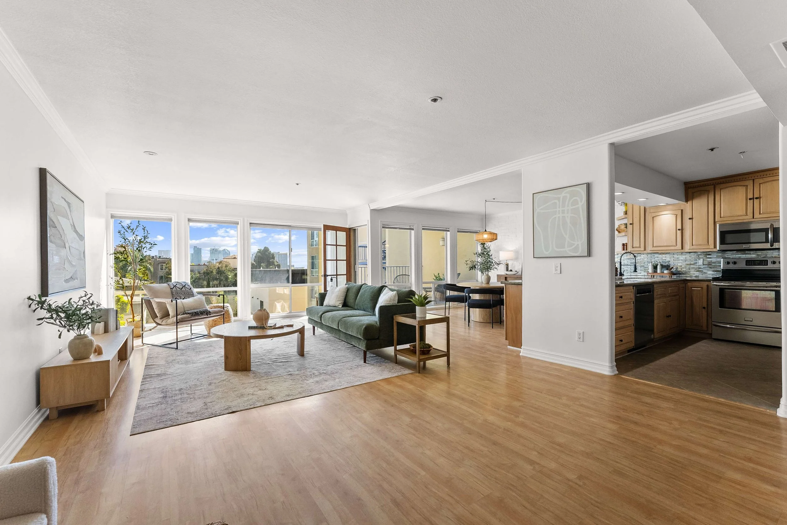 Spacious living room with large windows, light-colored walls, wooden floor, green sofa, coffee table, lounge chairs, and sliding glass doors leading to balcony. Open kitchen with wooden cabinets and stainless steel appliances visible in the background.