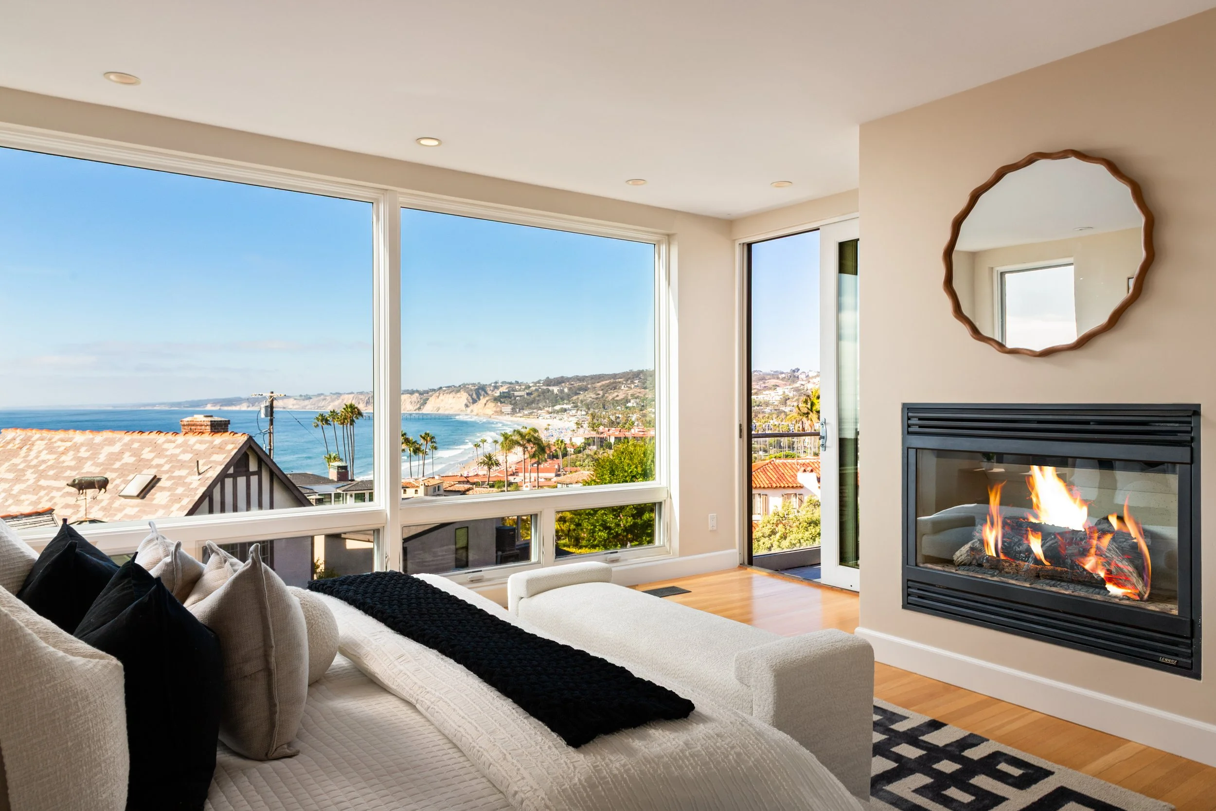 Living room with large windows showing a coastal view, a white couch with pillows, a fireplace with flames, and a round mirror on the wall.