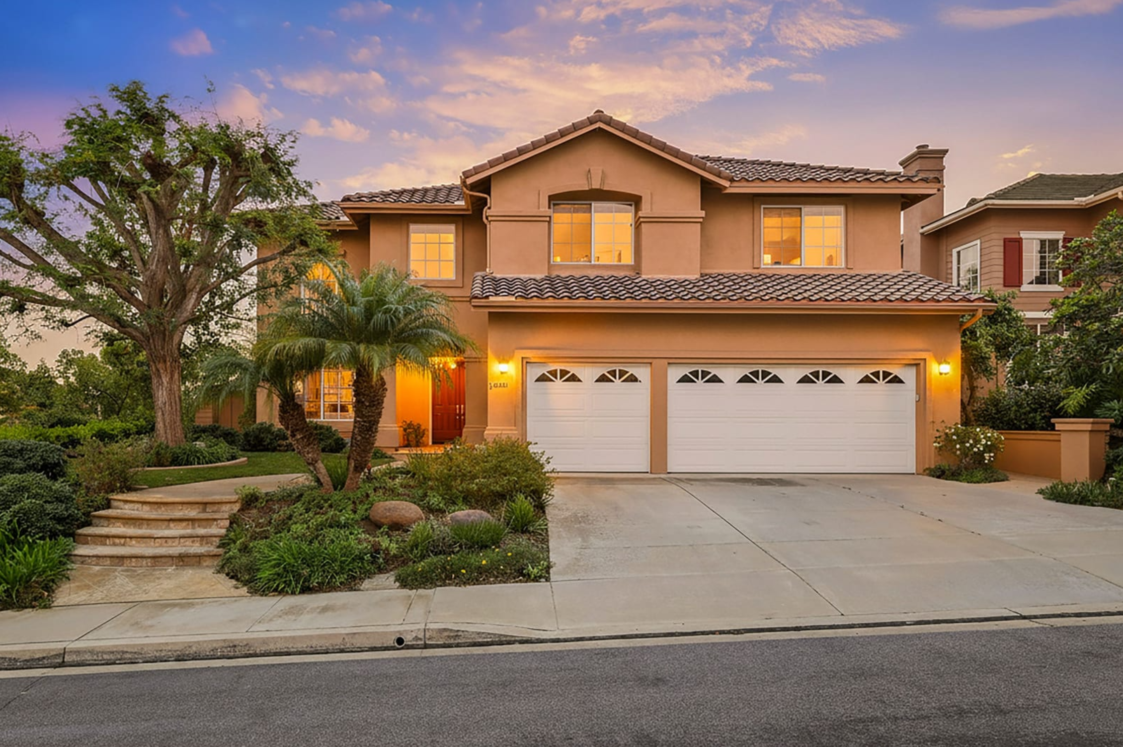 Two-story beige house with a tiled roof, illuminated by exterior lights, with a three-car garage, front yard with landscaped plants and trees, and a driveway at dusk.