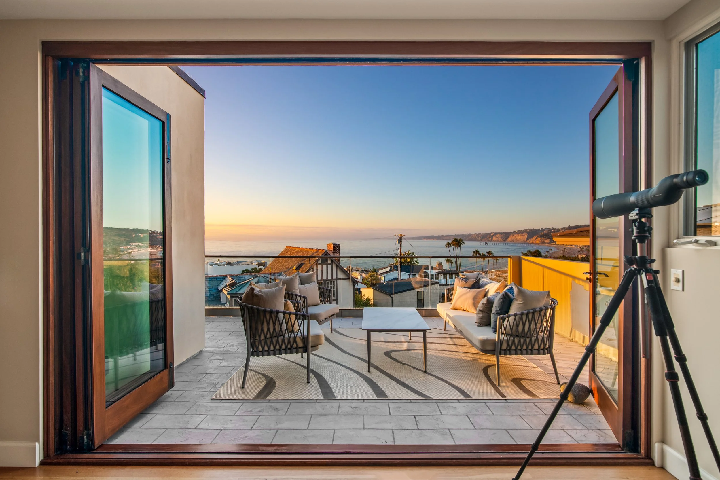 View from indoor to outdoor balcony with seating, overlooking ocean, houses, and palm trees at sunset.