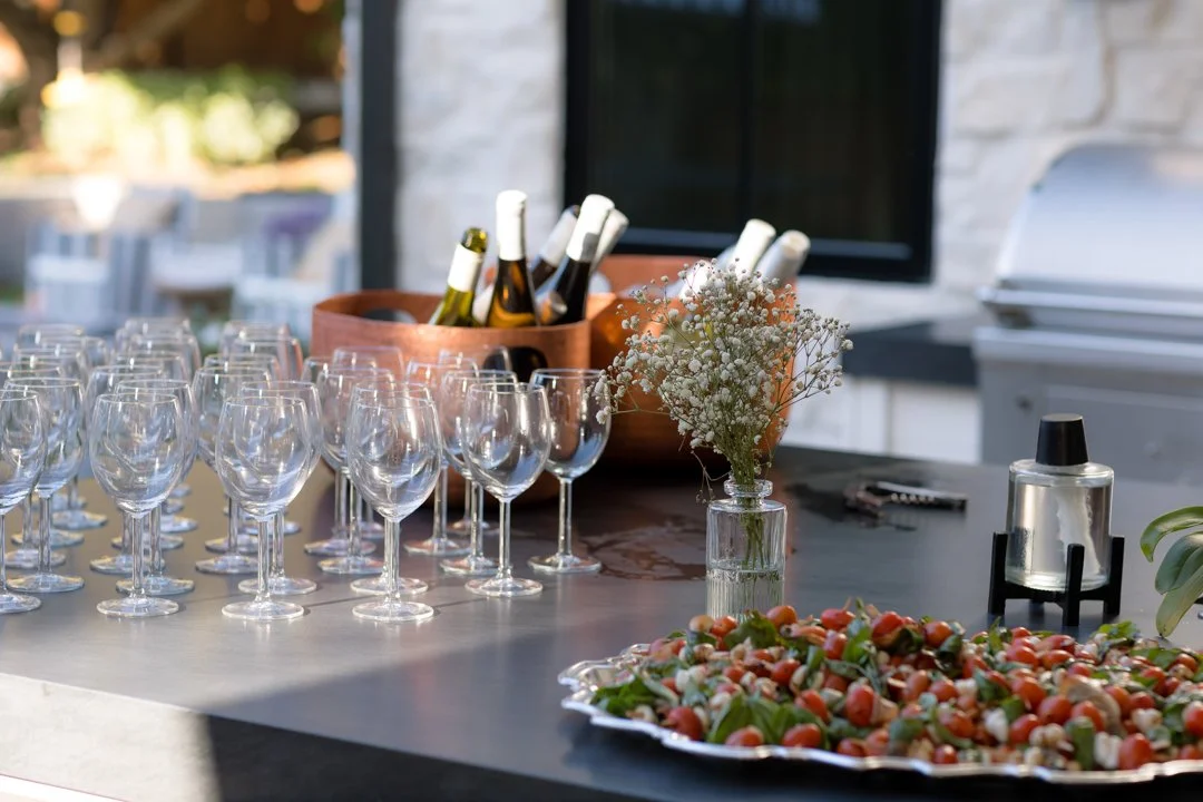 Empty wine and champagne glasses arranged on a table, with a vase of white baby's breath flowers, a plate of cherry tomatoes and basil, and bottles of wine in a wooden container, set outdoors near a stone fireplace.