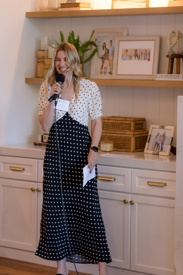 Woman smiling and speaking into a microphone in a cozy, decorated room with framed photographs and decor on shelves behind her.