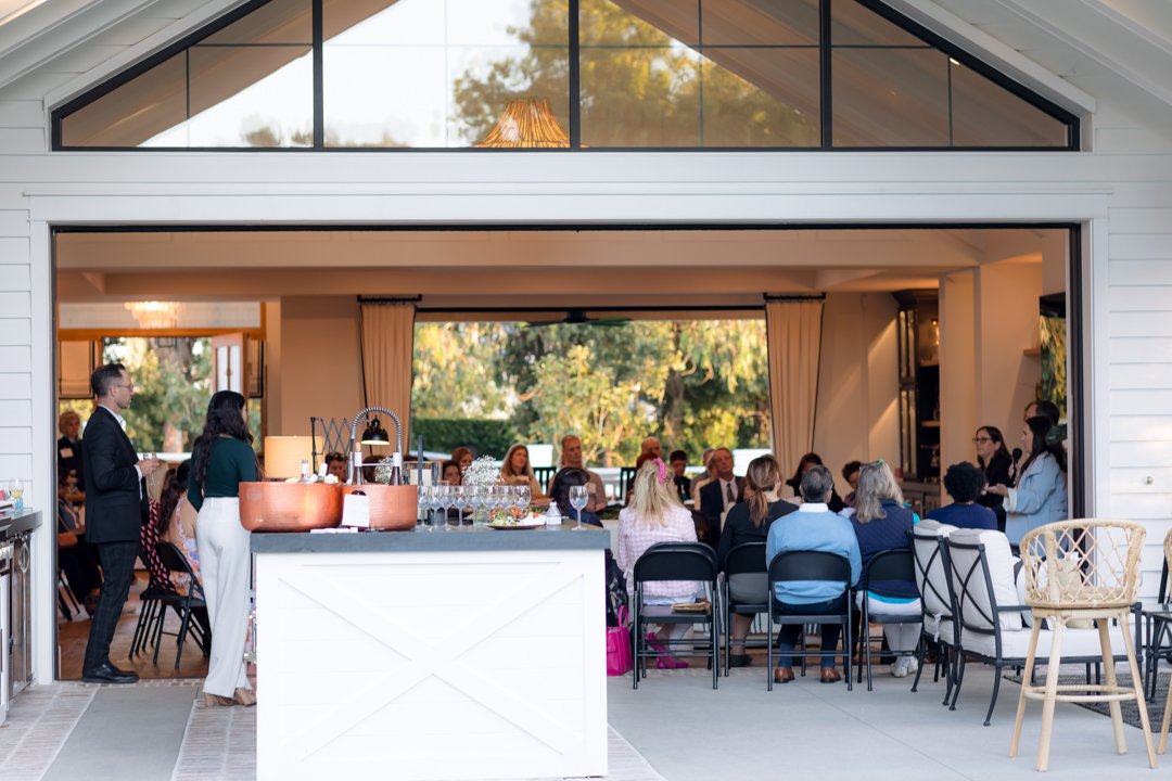 Guests attending a formal event or wedding reception dining inside a modern open-air venue with large windows and a view of trees outside.
