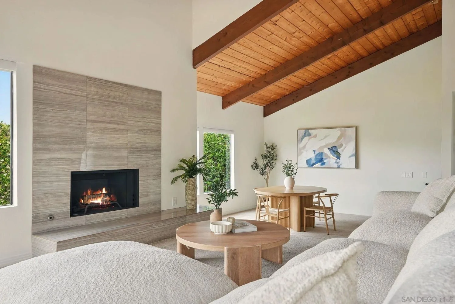 Living room with a fireplace, beige sofa, wooden round coffee table, two wooden chairs around a dining table, art on the wall, indoor plants, and a wooden ceiling with exposed beams.