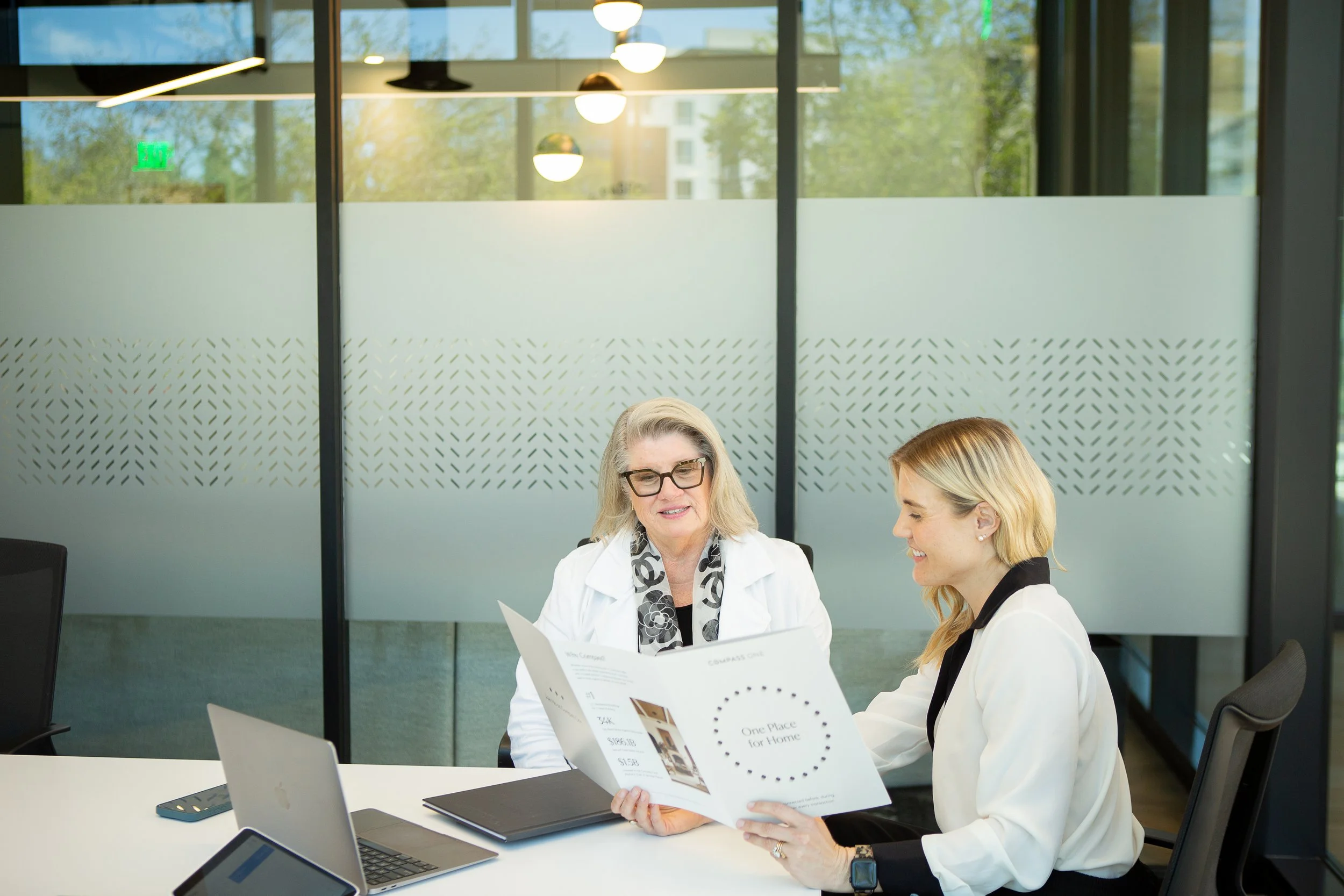 Two women sitting at a table in a modern office, looking at brochures and enjoying a conversation, with electronic devices on the table.