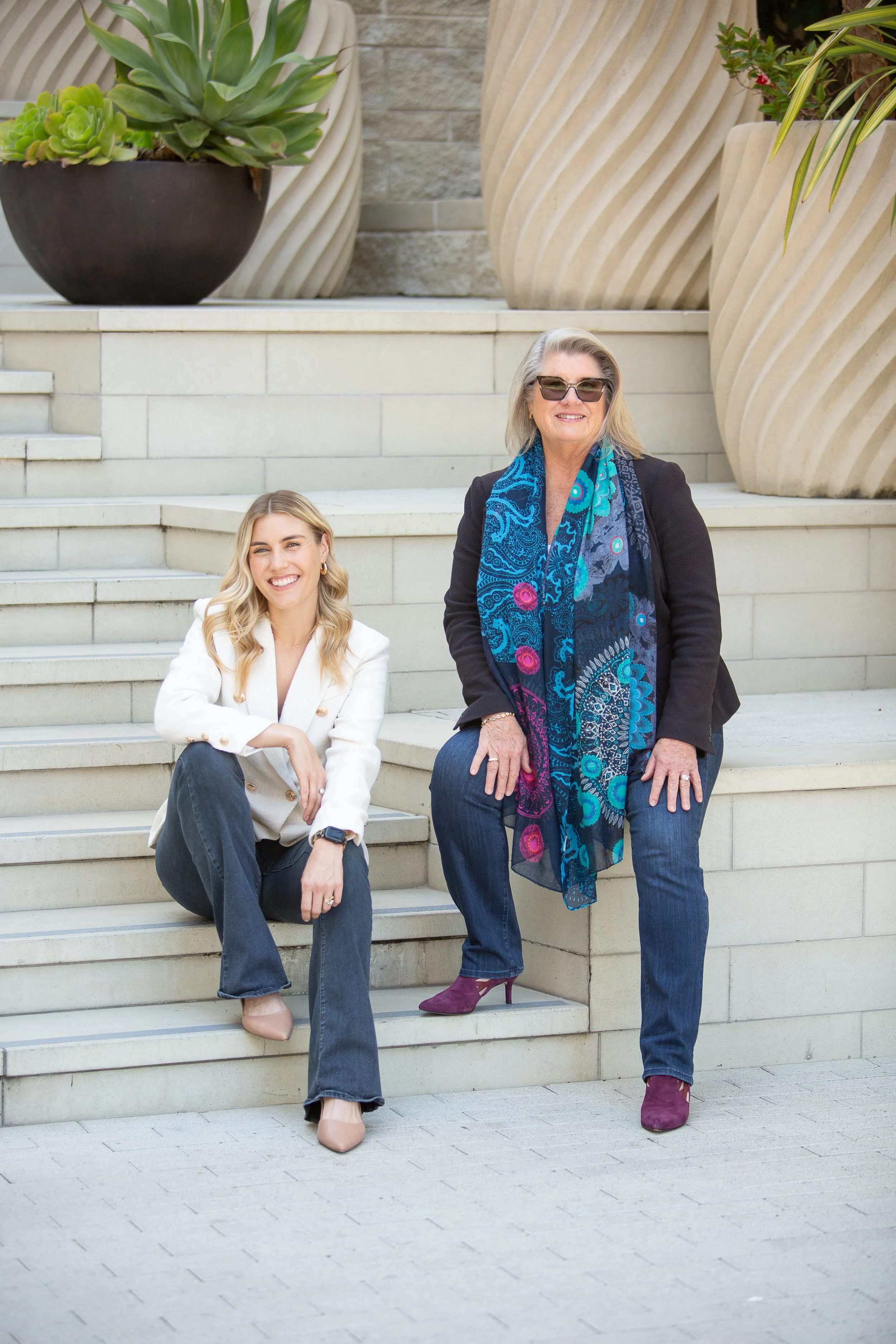 Two women sitting on marble steps outdoors, smiling. The younger woman has long blonde hair, is wearing a white blazer, dark jeans, beige heels, and a smartwatch. The older woman has shoulder-length gray hair, is wearing sunglasses, a black blazer, blue jeans, purple heels, and a vibrant blue patterned scarf.