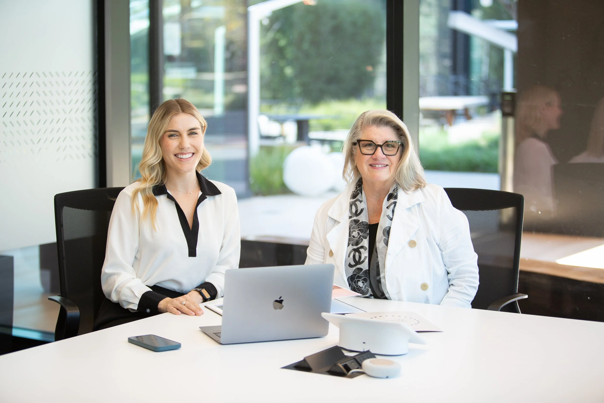 Two women sitting at a conference table in a modern office, smiling and looking at the camera, with a laptop and smartphone on the table.