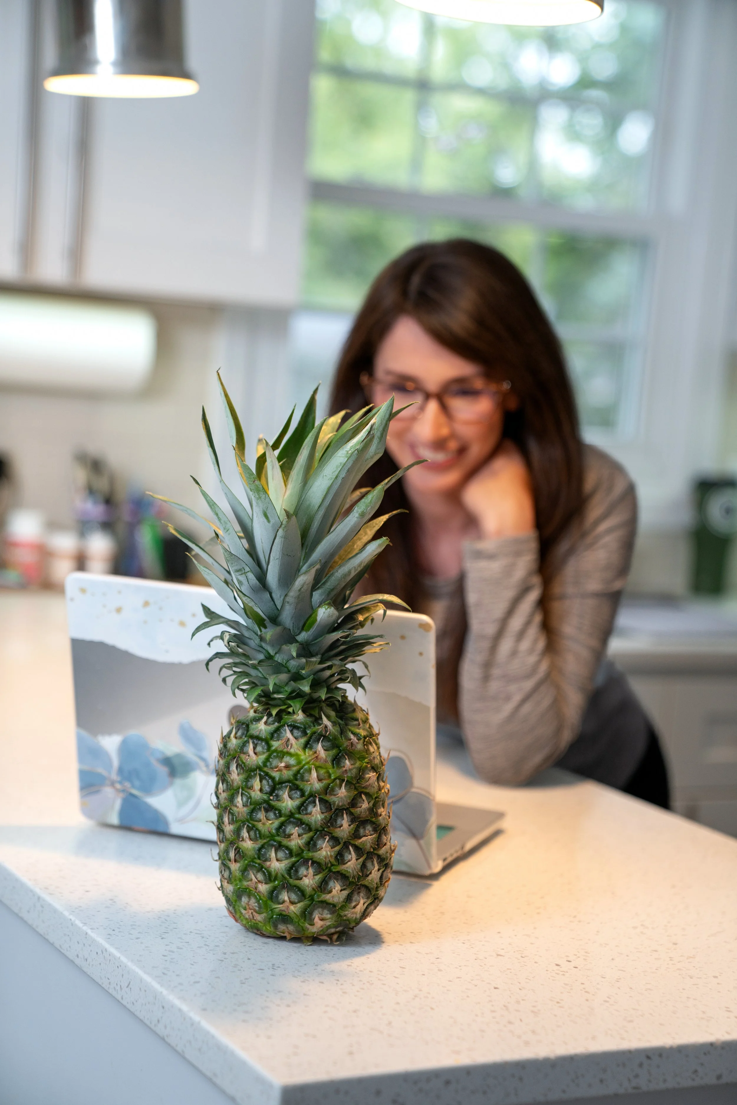 A woman with glasses and brown hair leaning on a white kitchen counter, smiling at a pineapple with its crown of green leaves prominently displayed in the foreground.